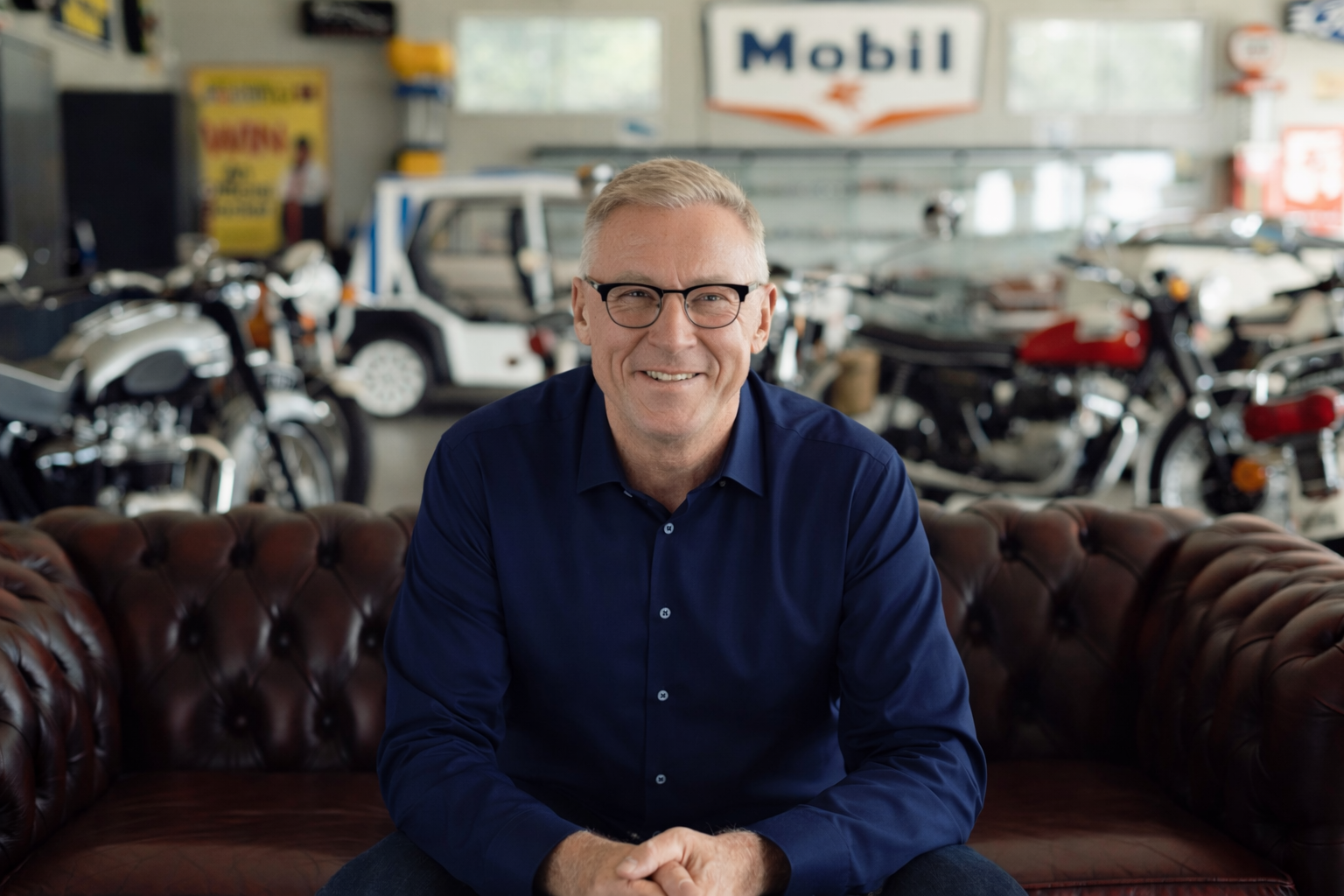 A smiling man with glasses and gray hair sitting on a brown leather couch in a vintage car and motorcycle showroom.