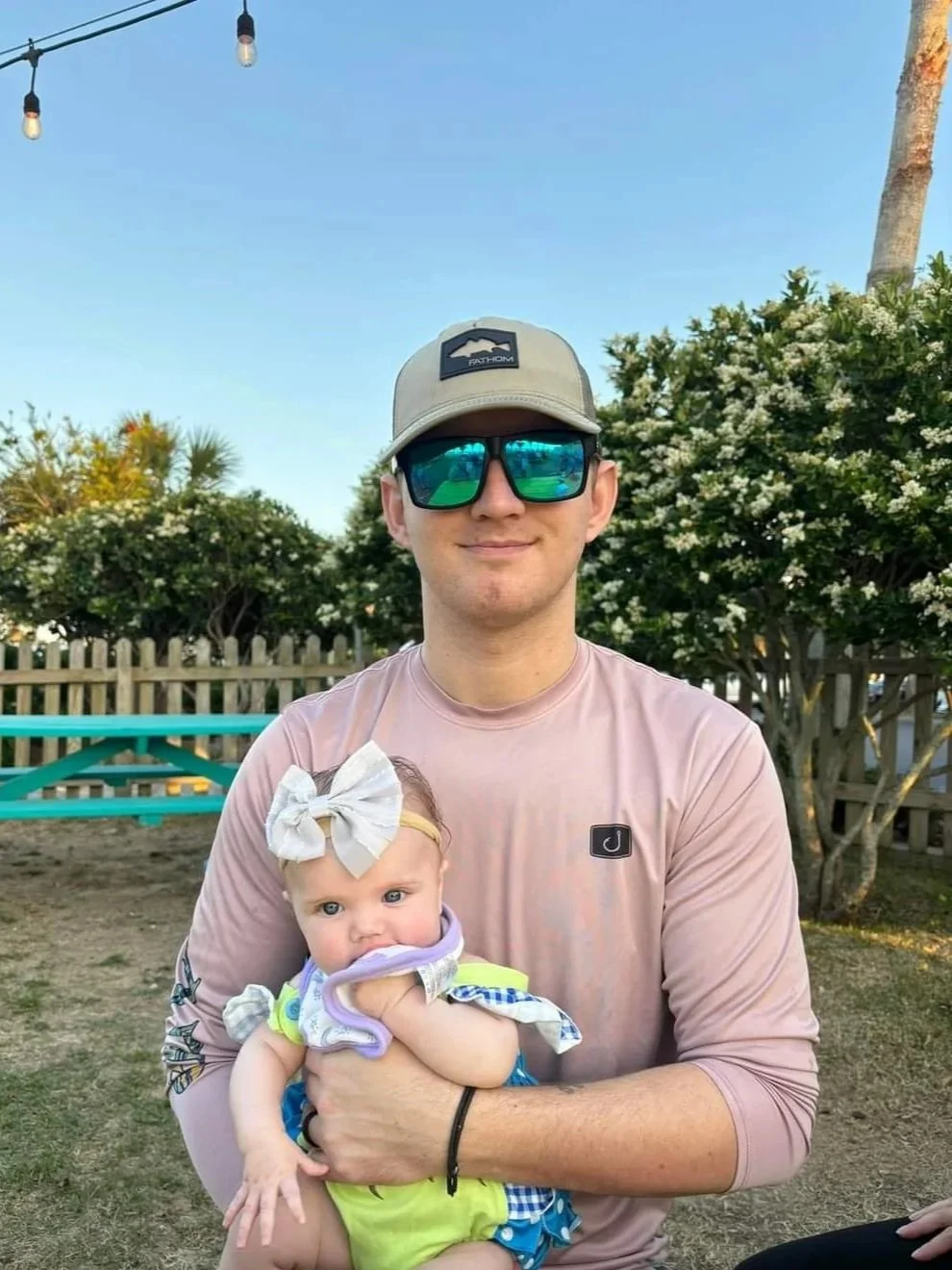 A man wearing sunglasses, a beige cap, and a pink long-sleeve shirt is holding a baby with a white bow headband and colorful clothes outdoors next to a wooden fence and bushes.