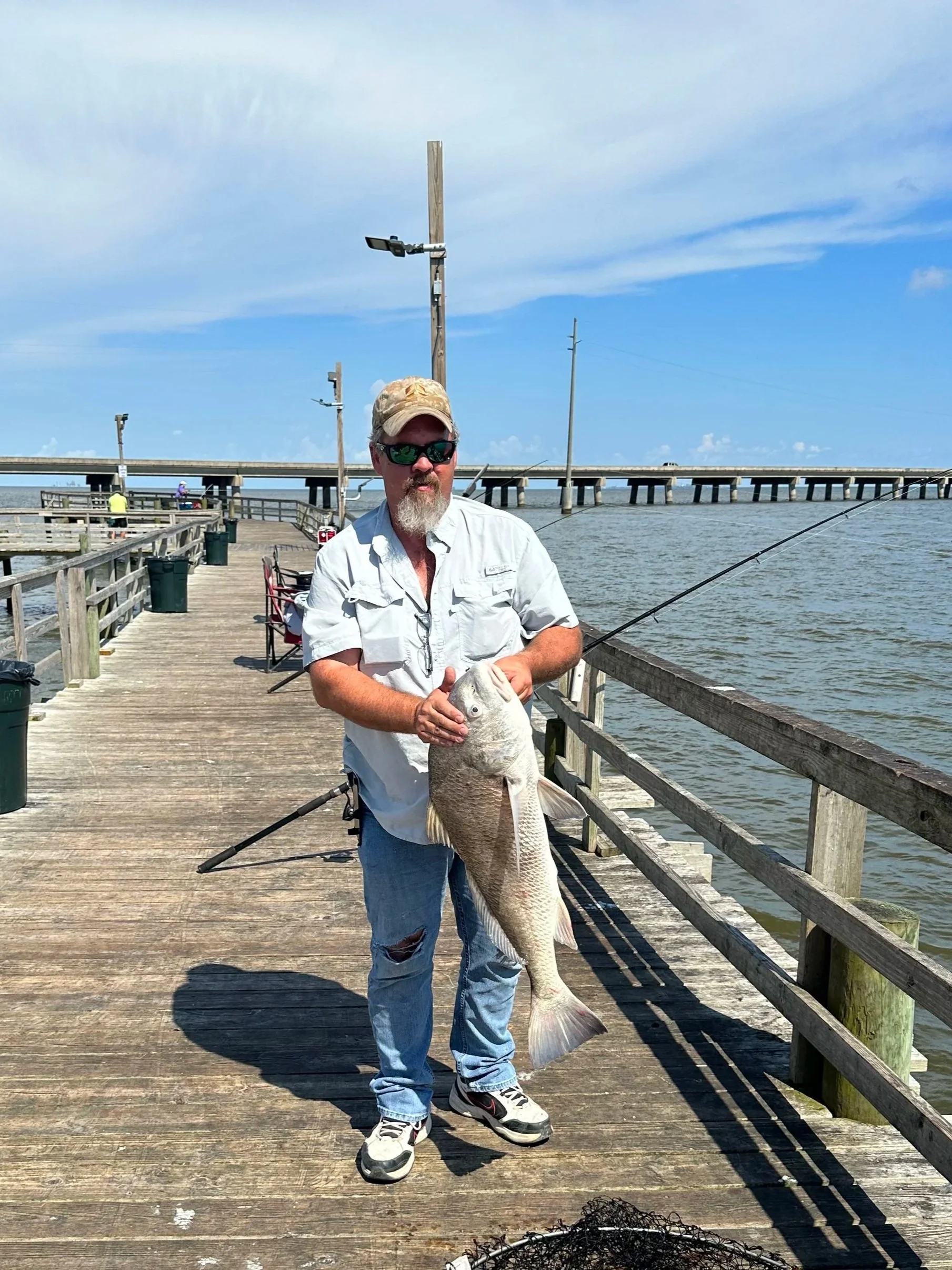 A man with a beard wearing sunglasses, a beige cap, a white short-sleeved shirt, ripped jeans, and sneakers, holding a large fish on a wooden pier over a body of water, with fishing rods, some people, and a bridge in the background under a blue sky.