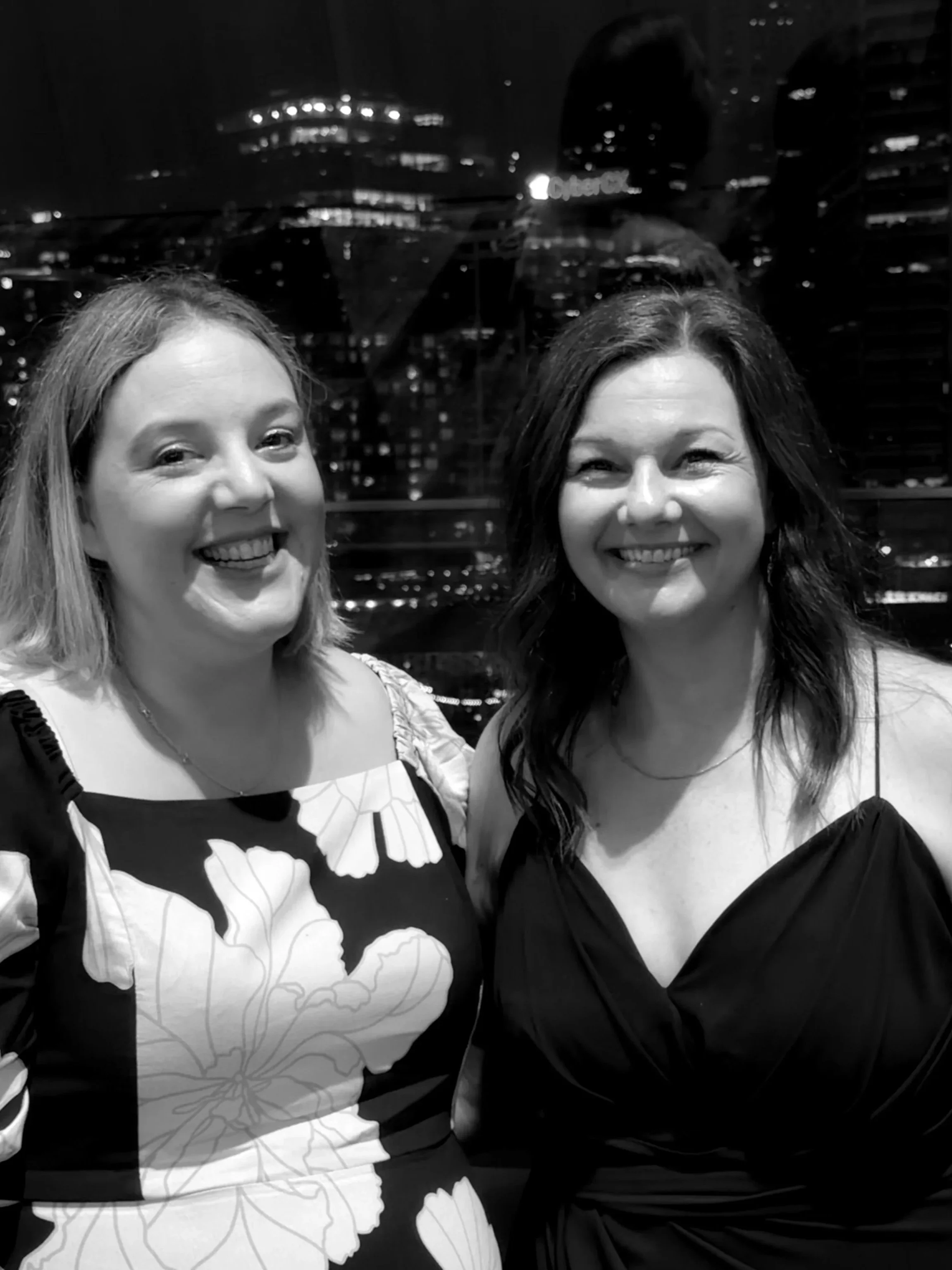 Two women smiling at night in front of a cityscape with tall buildings and lights.