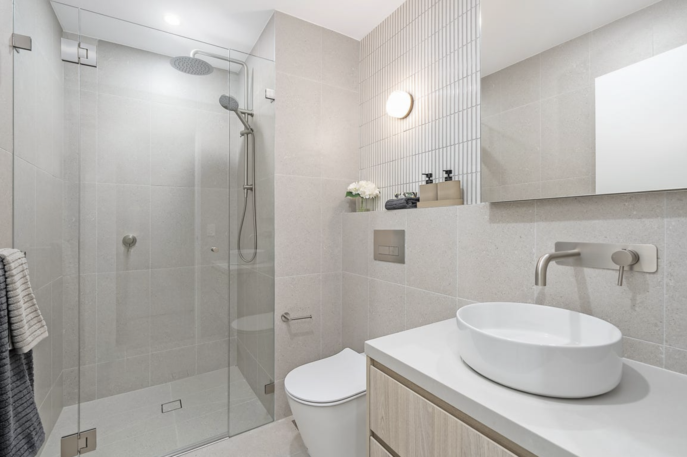 Modern bathroom with a glass-enclosed walk-in shower, a wall-mounted toilet, a white vessel sink on a wooden vanity, a large mirror, and minimalist decor.