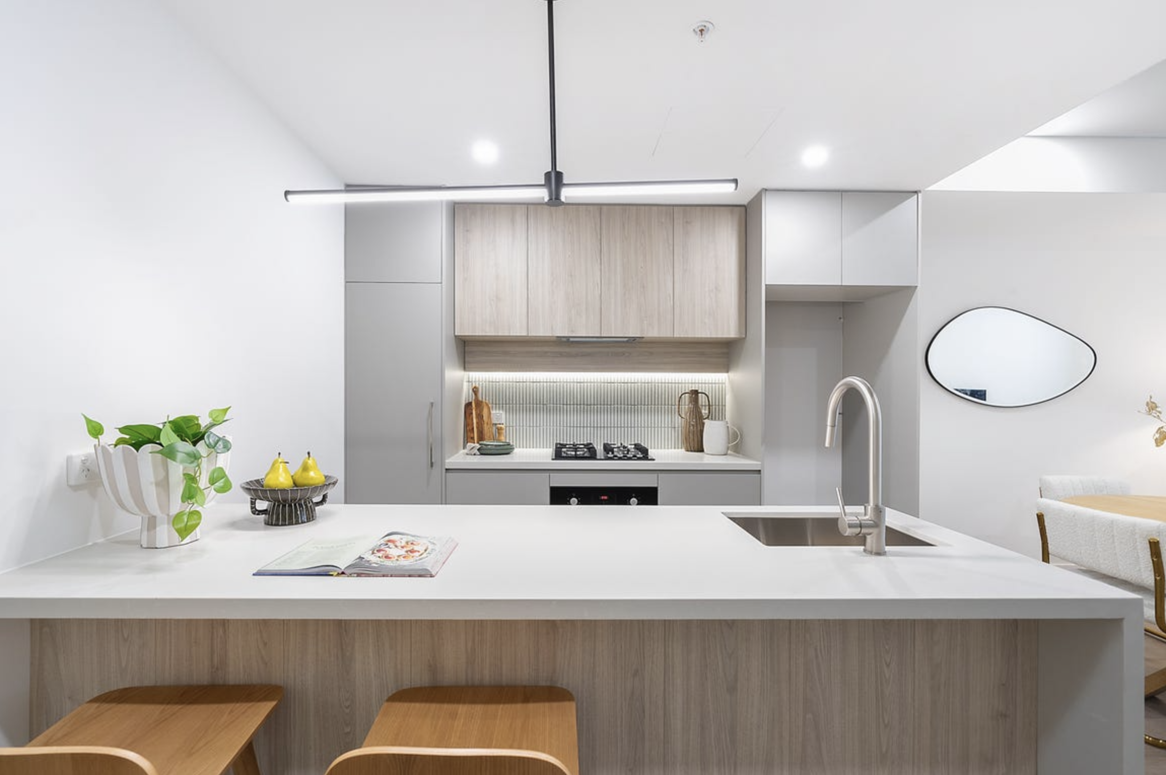 Modern kitchen with white and wood cabinets, a white countertop island with a sink, potted plant, decorative pears, open cookbook, and a dining area with wooden chairs and an oval mirror on the wall.