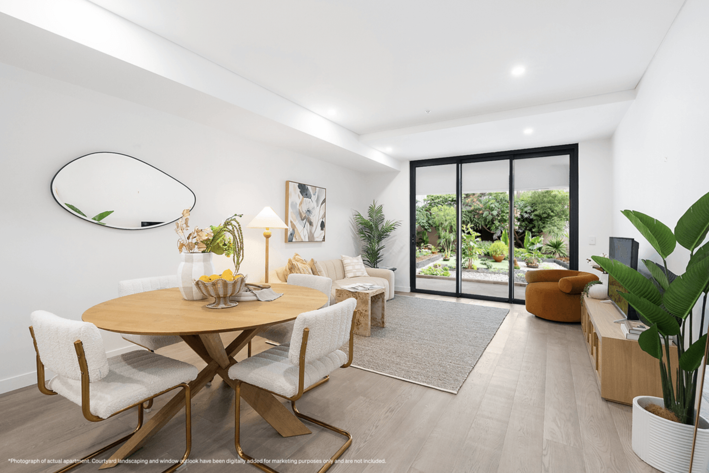Modern living room with white walls, wooden flooring, a round dining table with four chairs, a beige sofa, a round brown armchair, a TV, large sliding glass doors opening to a lush garden, and various indoor plants.