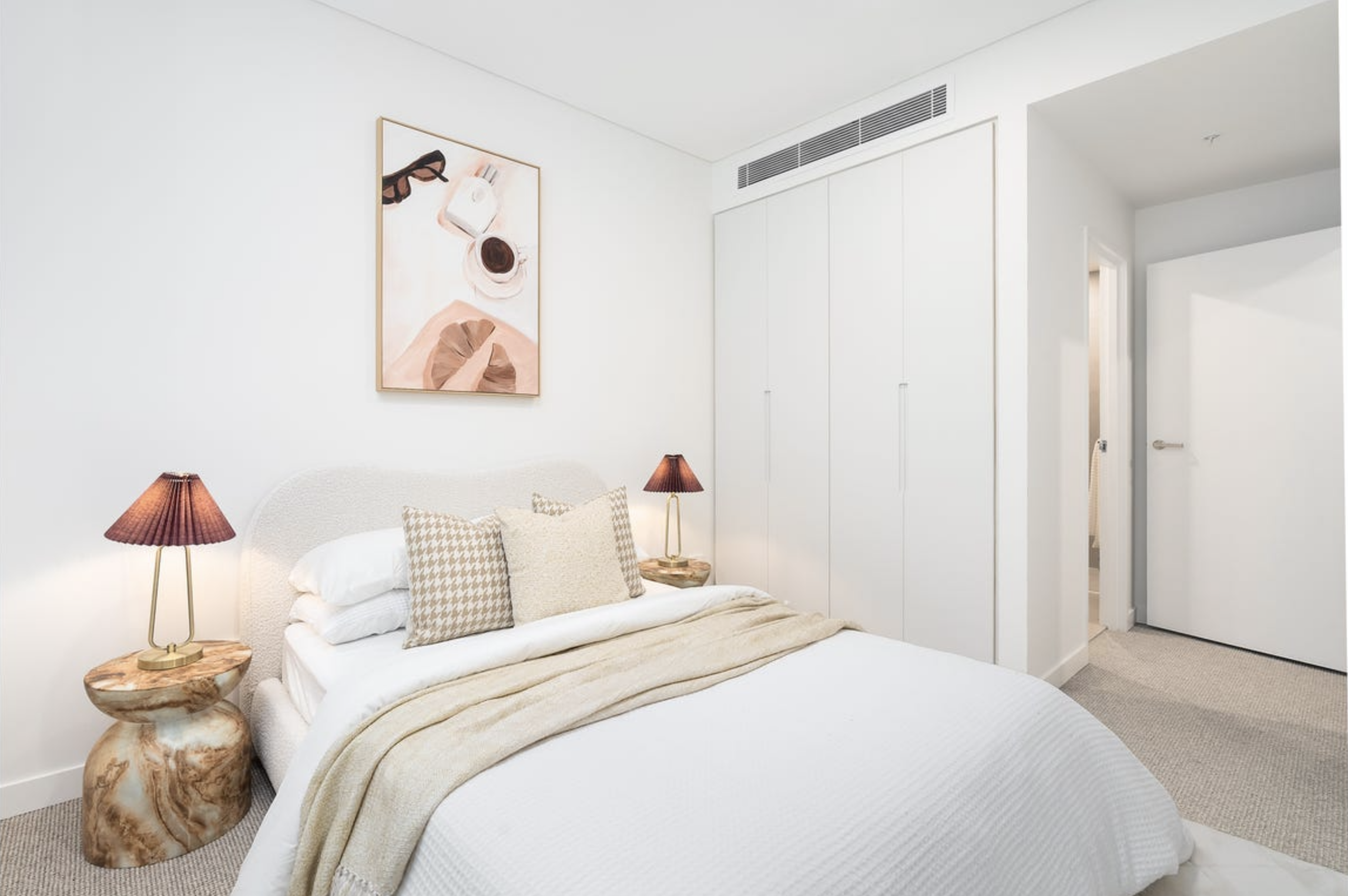 A neatly made white bedroom with a bed featuring beige pillows and a cream blanket, two wooden side tables with red lamps, a framed abstract art piece on the wall, white closet doors, and a partially open door to another room.