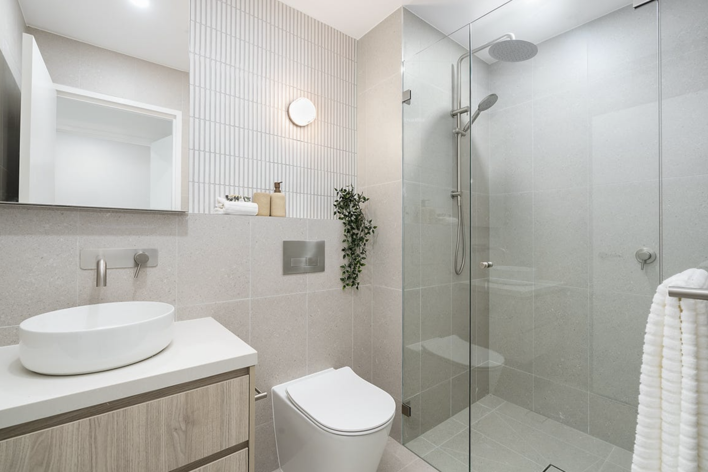 Modern bathroom with beige tile walls and floor, a glass-enclosed shower with rainfall and handheld showerheads, a wall-mounted toilet, a light wood vanity with a vessel sink, a mirror, a small potted plant, towels, and minimalist decor.
