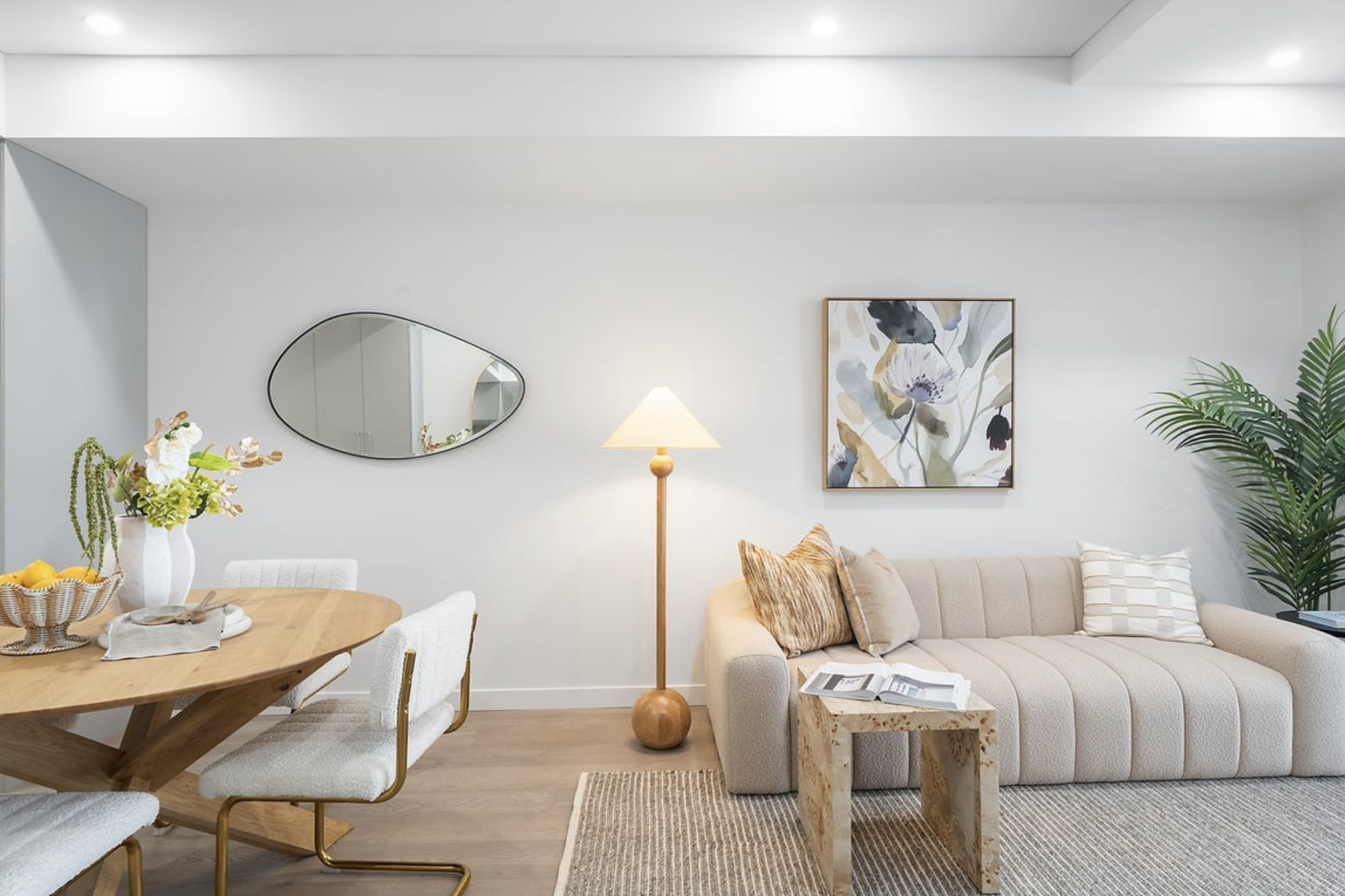 Living room with beige sofa, floral artwork, lamp, and potted plant; dining table with chairs, flowers, and lemons.