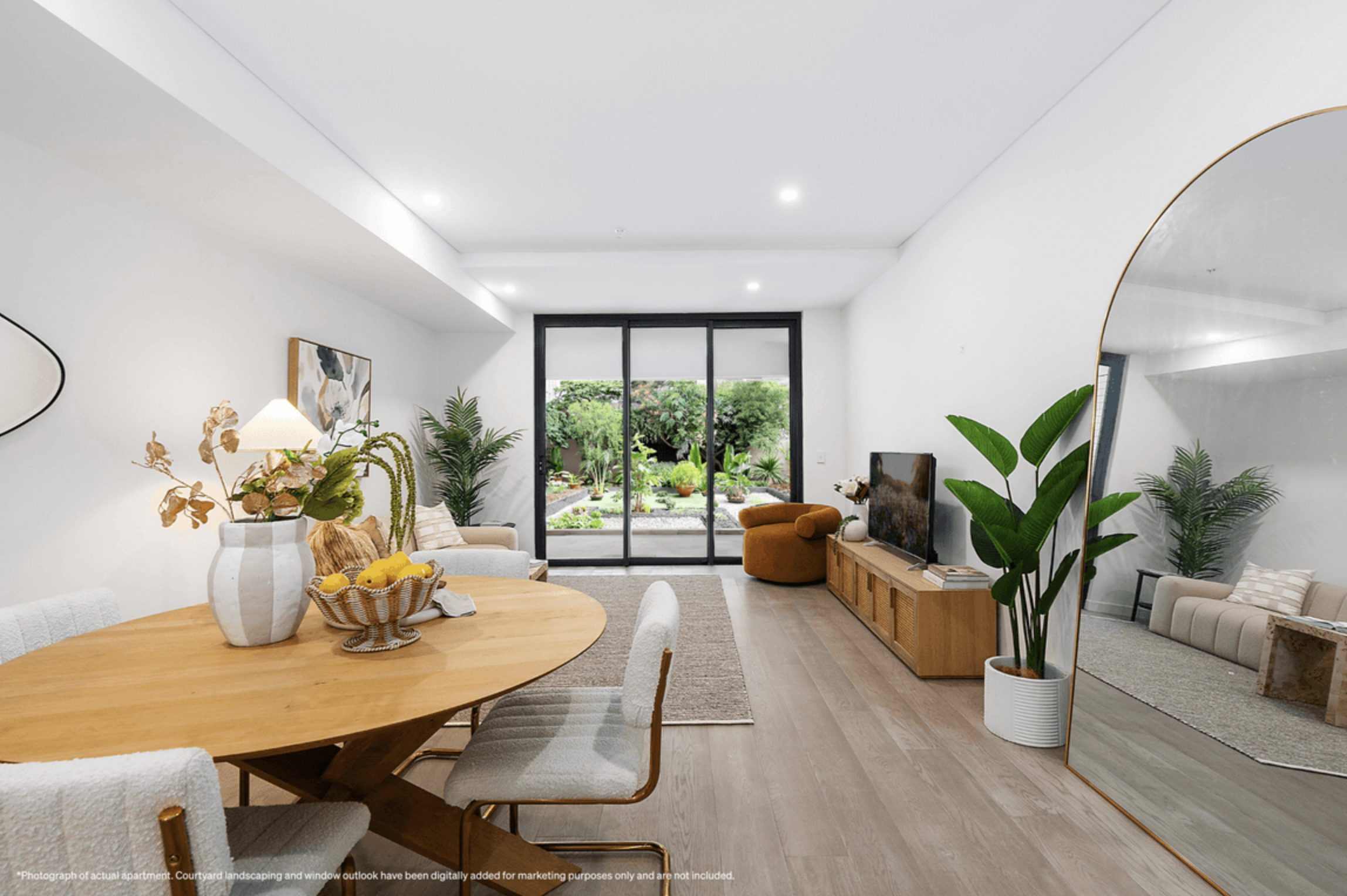 Modern living room with a wood dining table, surrounded by four upholstered chairs, a white sofa with pillows, a media console with a TV, potted plants, and a sliding glass door leading to a garden.