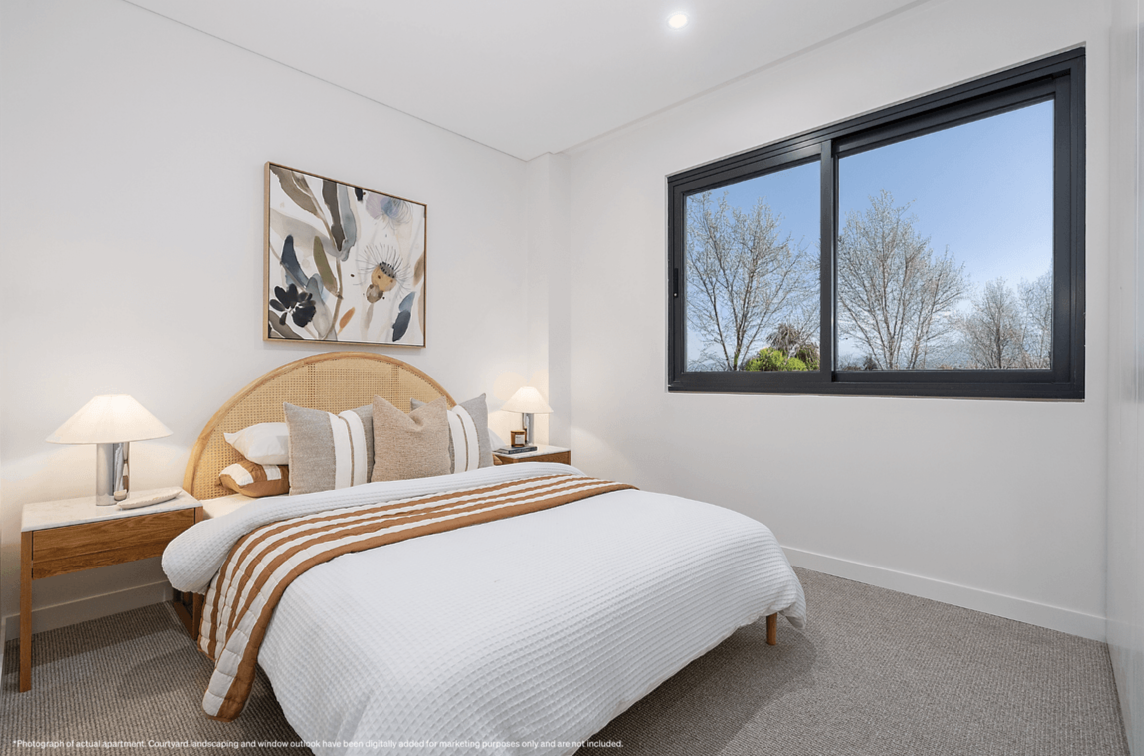 Bedroom with white walls, beige carpet, a bed with a white quilt and brown striped throws, multiple decorative pillows, a wooden headboard, and two bedside tables with lamps. There is a large window with black framing showing a view of leafless trees