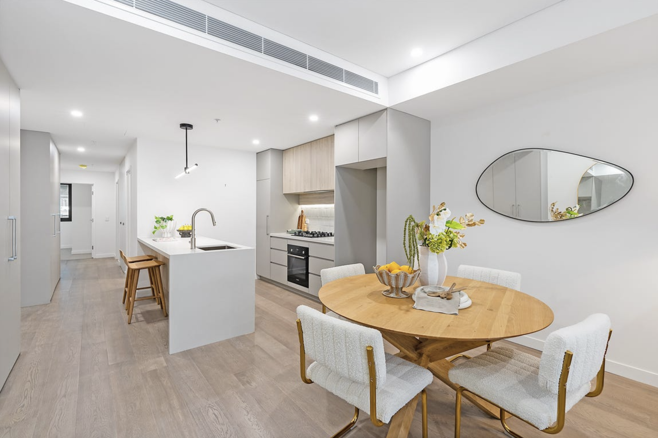 Modern open-concept kitchen and dining area with white walls, wooden flooring, a round wooden dining table with four white upholstered chairs, a kitchen island with a sink and wooden stools, light gray cabinetry, a gas stove, and minimal decorative e