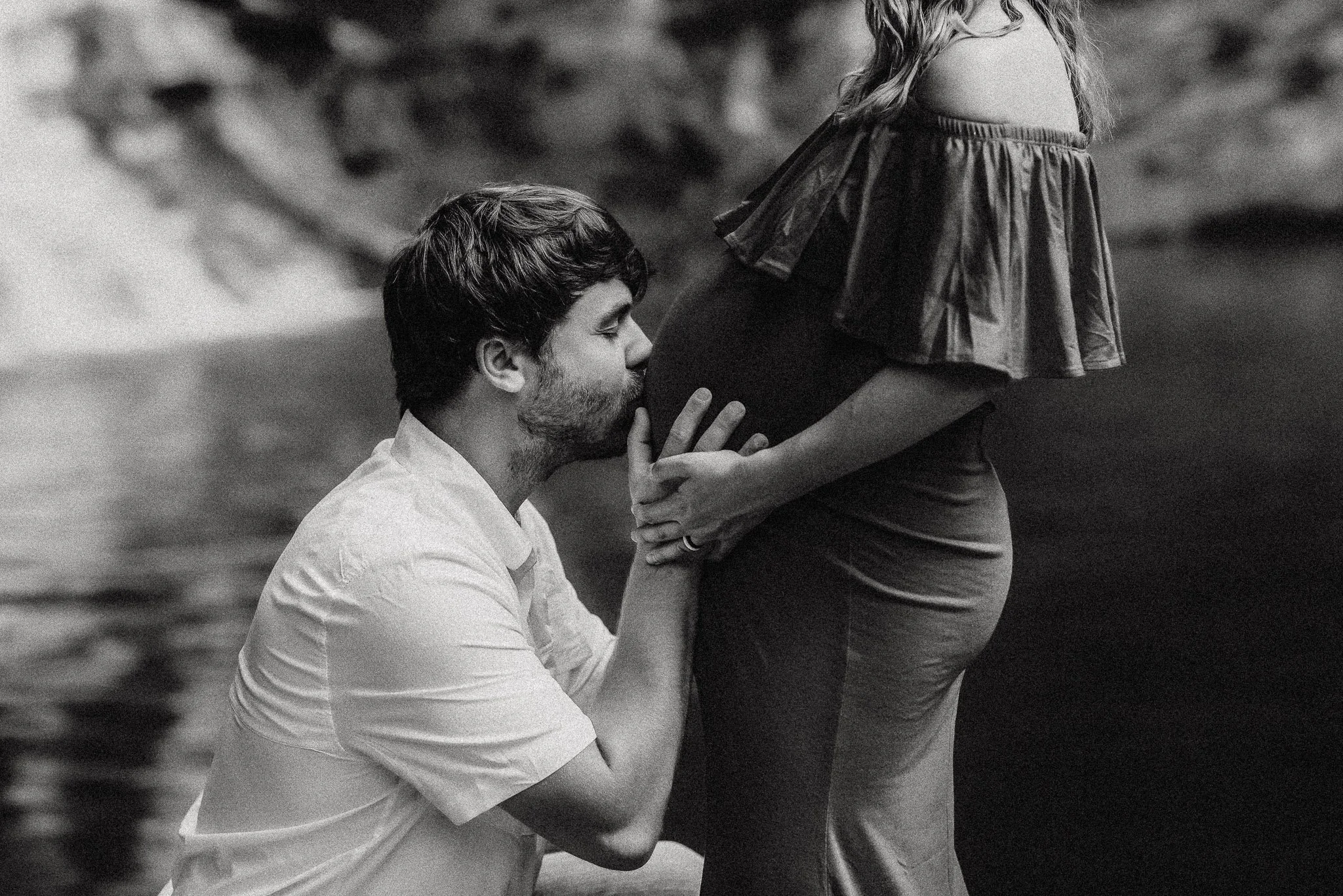 Black and white emotional image of husband kissing pregnant wife belly maternity session at Hightower Falls