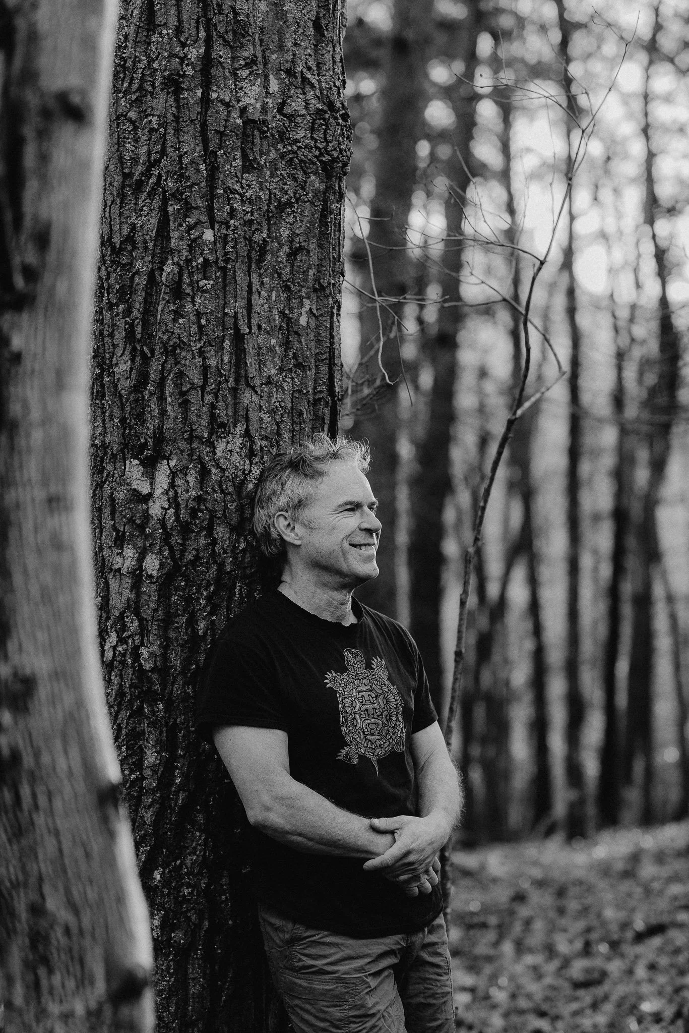 Black and white image of man smiling while leaning on tree in the woods portrait session in Rydal, Georgia