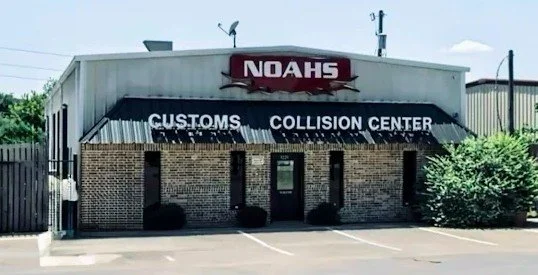 Front view of a building labeled NOAHS, with signs for Customs and Collision Center, situated in a parking lot with bushes outside.