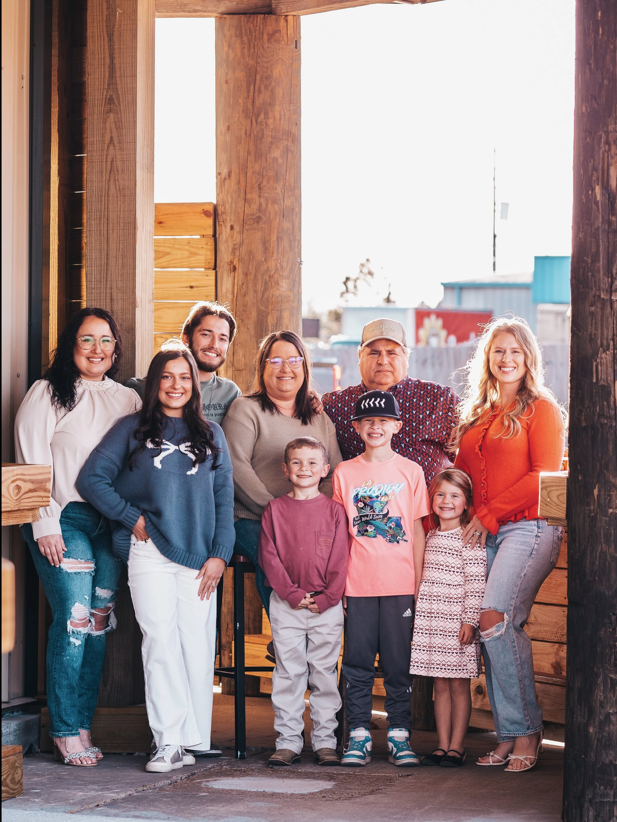 A group of ten people, including children and adults, posing together outdoors in front of a partially constructed wooden structure during daytime. They are smiling and standing close together, dressed casually.