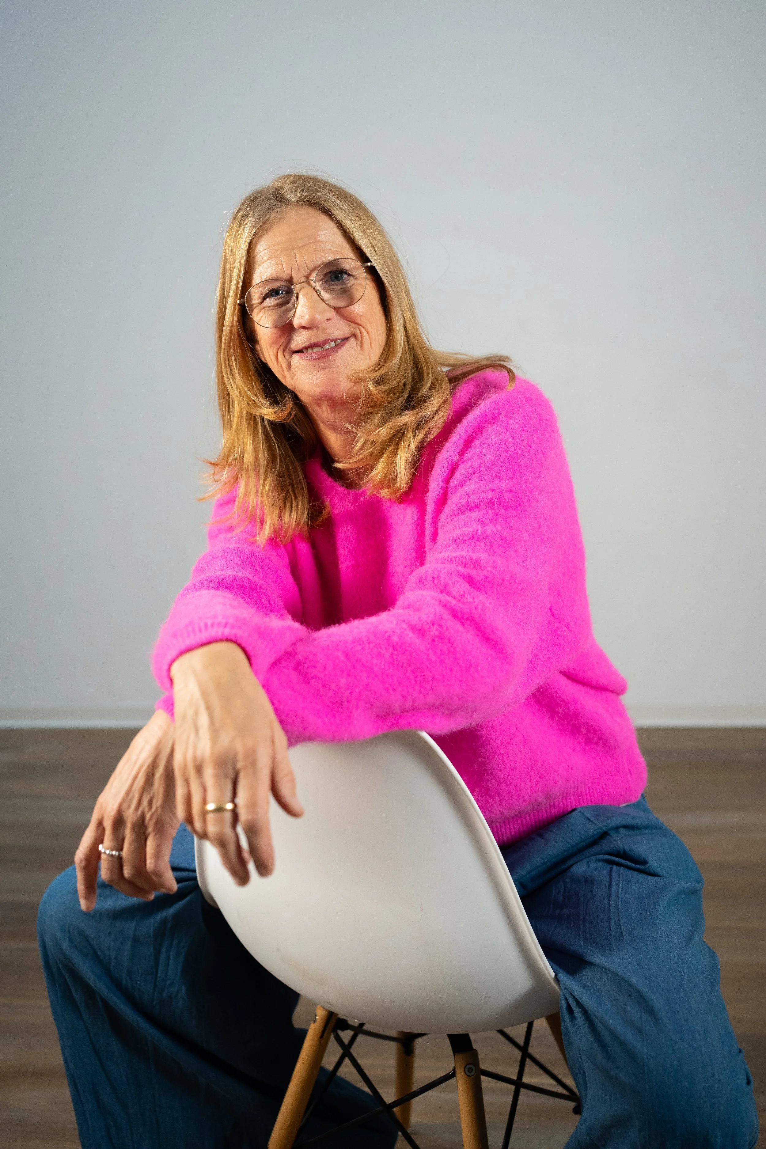 Eine ältere Frau in einer pinken Pullover sitzt auf einem weißen Stuhl, lächelt und sieht freundlich aus.