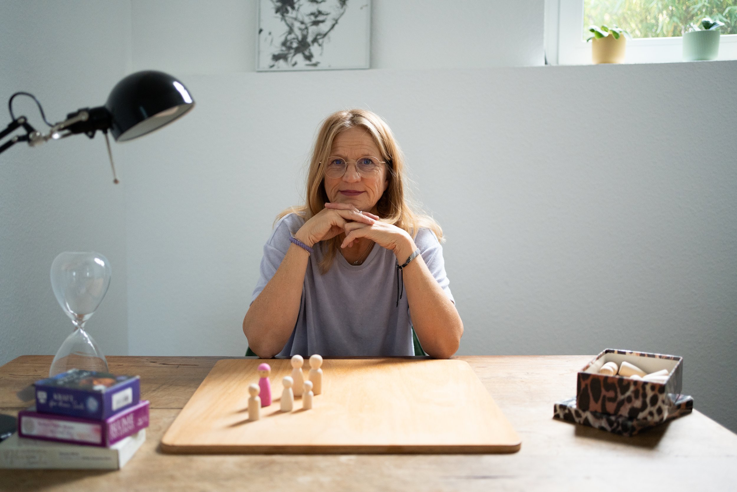 Eine Frau sitzt an einem Holztisch mit Brettspiel, vor ihr stehen kleine Spielfiguren. Es gibt eine Tabelle mit Abdeckungen, eine Leuchtlampe, eine Sanduhr, und auf der rechten Seite eine dekorative Schachtel mit Spielsteinen. Im Hintergrund ein Fenster mit Pflanzen auf der Fensterbank und ein Kunstwerk an der Wand.