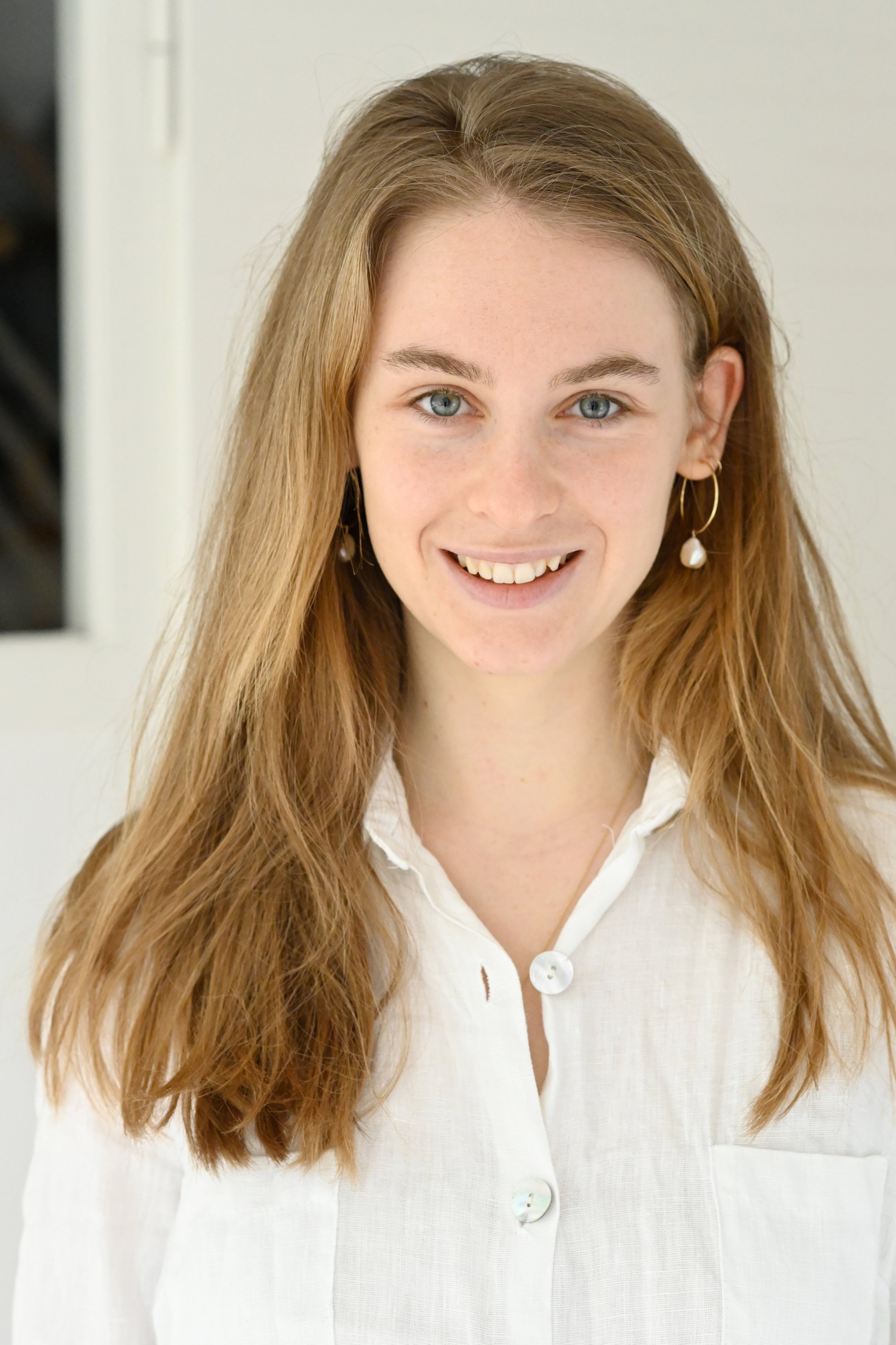 Ein junges Mädchen mit langen, hellbraunen Haaren, blaue Augen und freundlichem Lächeln, trägt eine weiße Bluse und goldene Ohrringe.