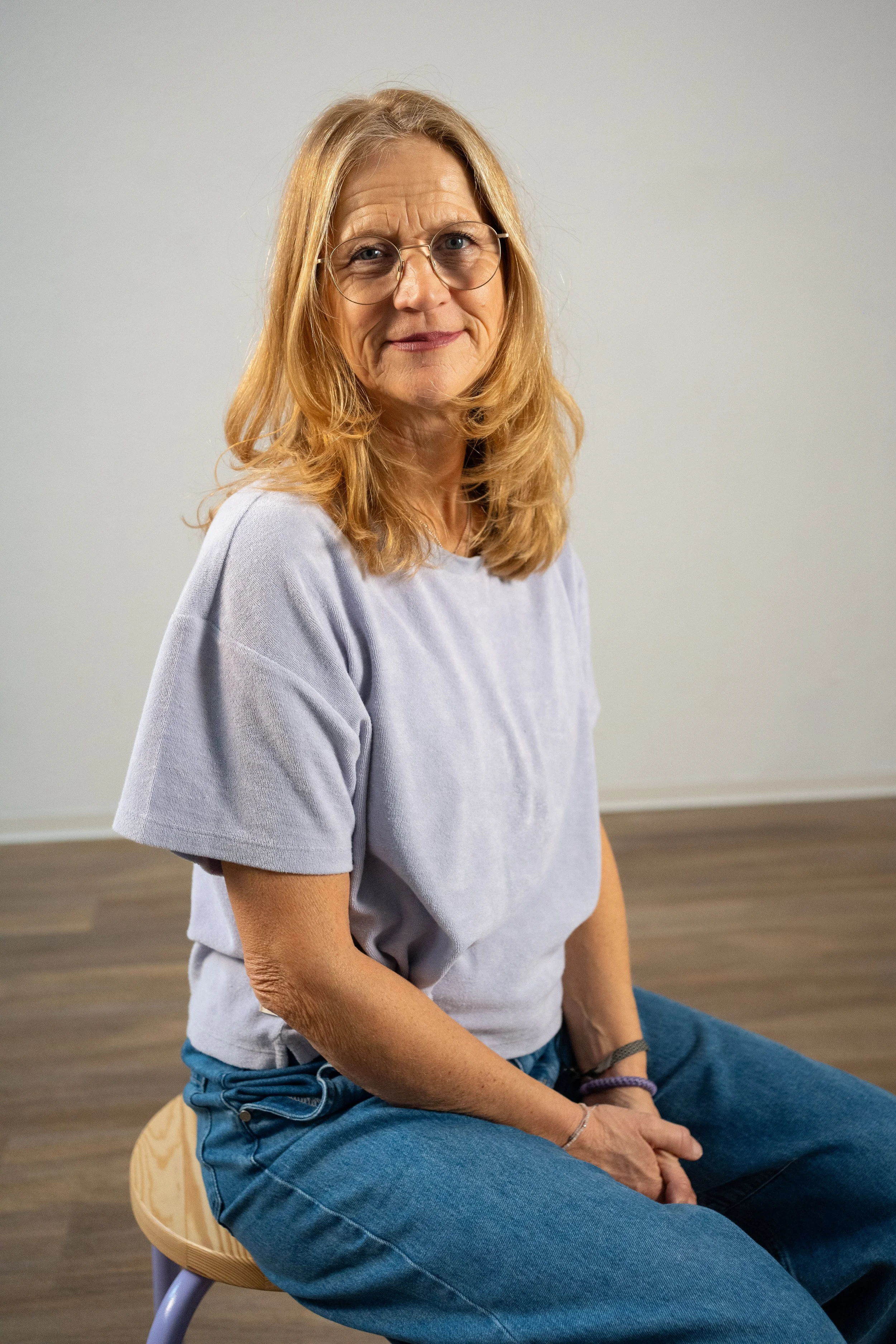 Eine Frau mit Brille sitzt auf einem Hocker vor weißer Wand, trägt ein lilafarbenes T-Shirt und blaue Jeans.