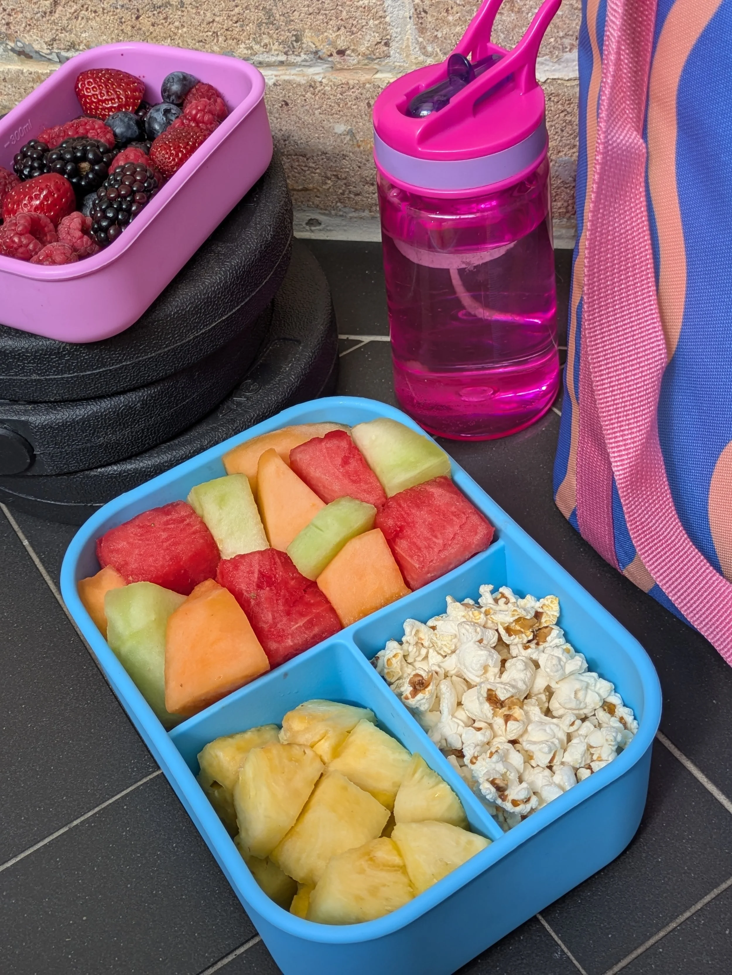 A plastic blue divided container filled with various cut melons including watermelon, cantaloupe, honey dew, pineapple, and watermelon, placed on a dark tiled floor next to a pink water bottle and a striped pink and blue fabric bag, with a brick wall background.
