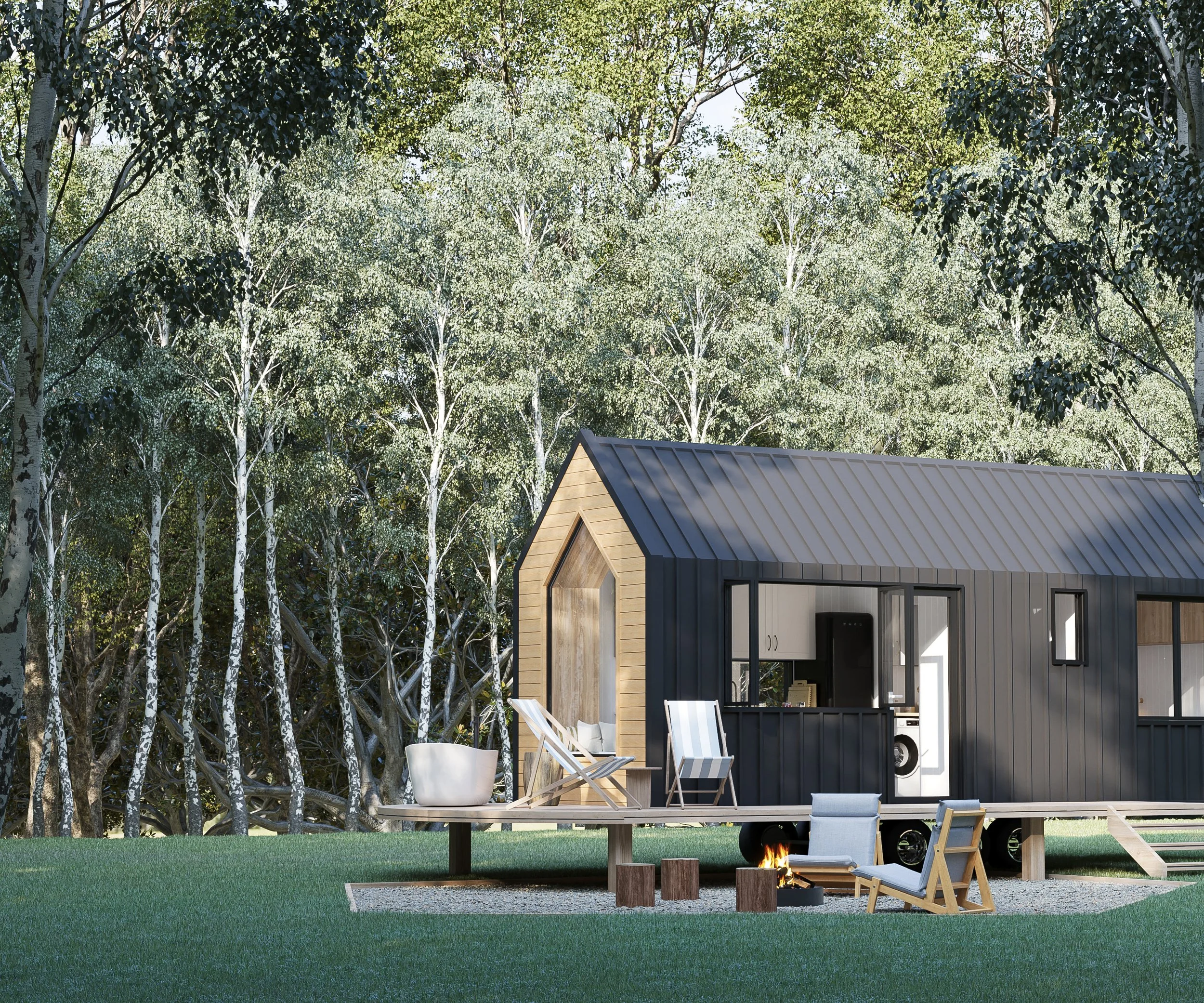 A tiny black and wood cabin on wheels in a grassy clearing with trees in the background. There is an outdoor seating area with chairs and a fire pit.
