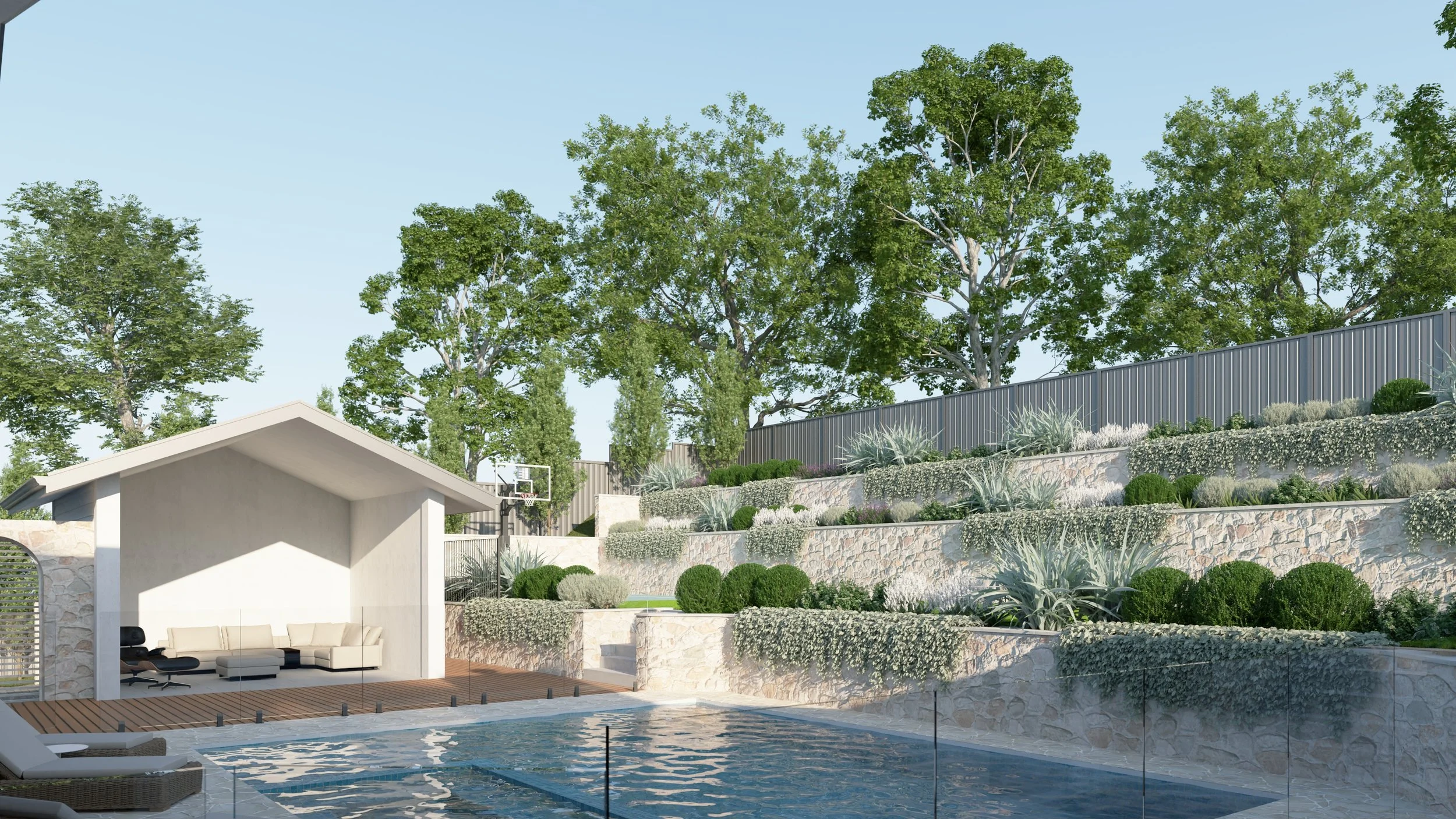 A backyard with a swimming pool, a white poolside lounge area with a sofa and chairs under a small shelter, surrounded by tiered stone and plant landscaping, tall trees, and a grey fence in the background.