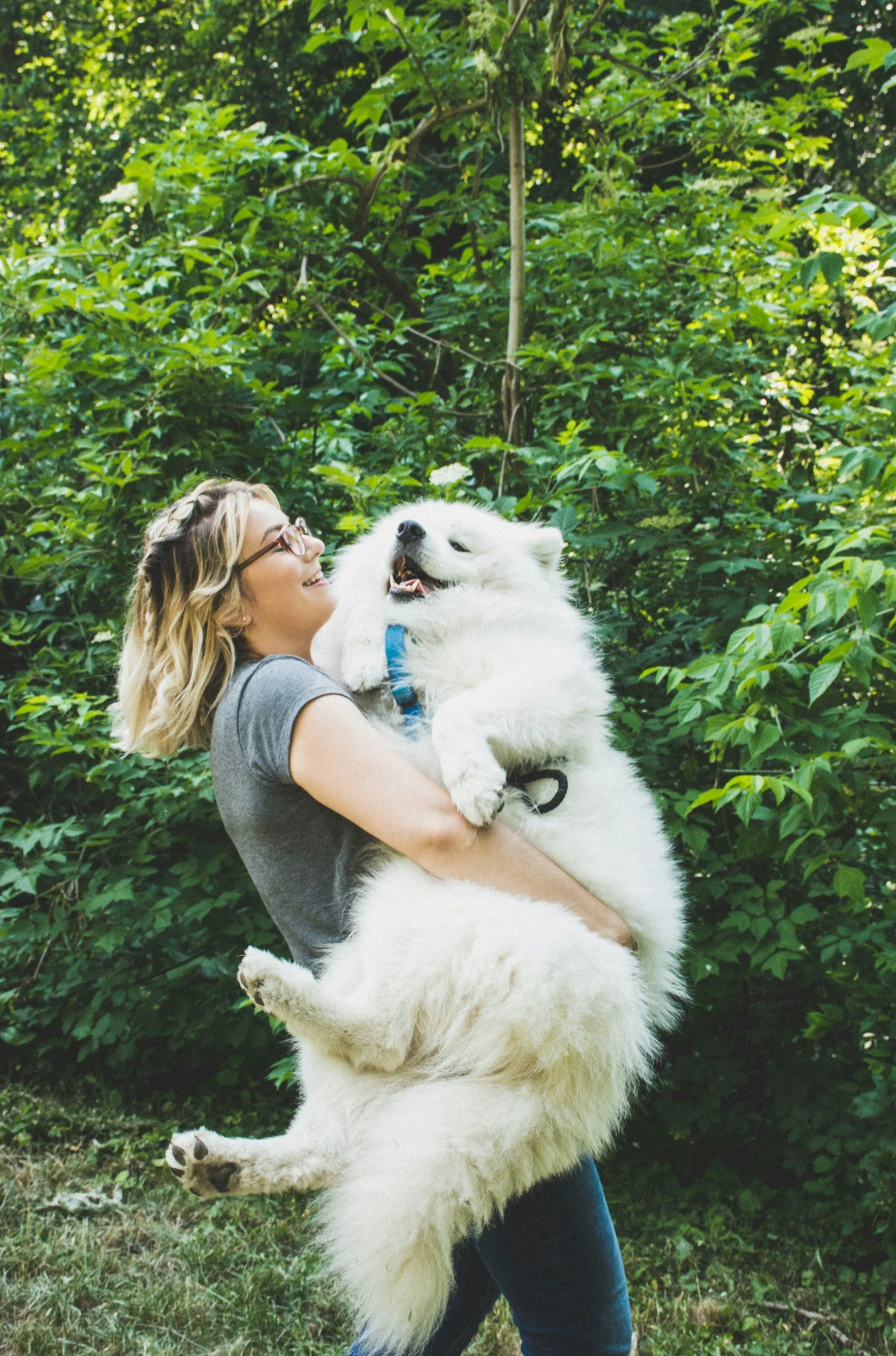 A woman with glasses and blonde hair holding a large, fluffy white dog in a green, wooded outdoor setting.