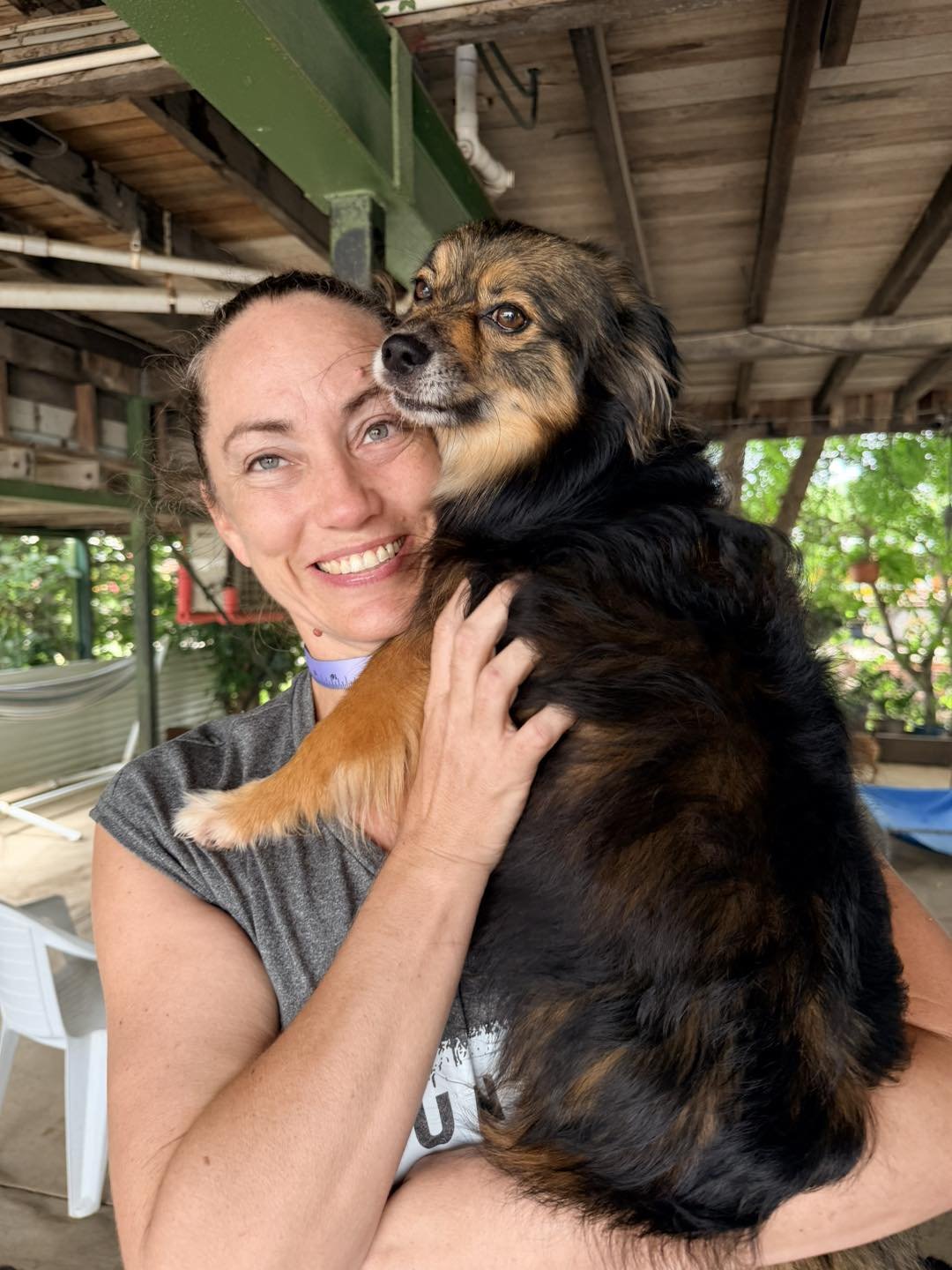 A woman smiling and holding a large, fluffy dog with black, brown, and tan fur under a wooden roofed outdoor area with greenery in the background.
