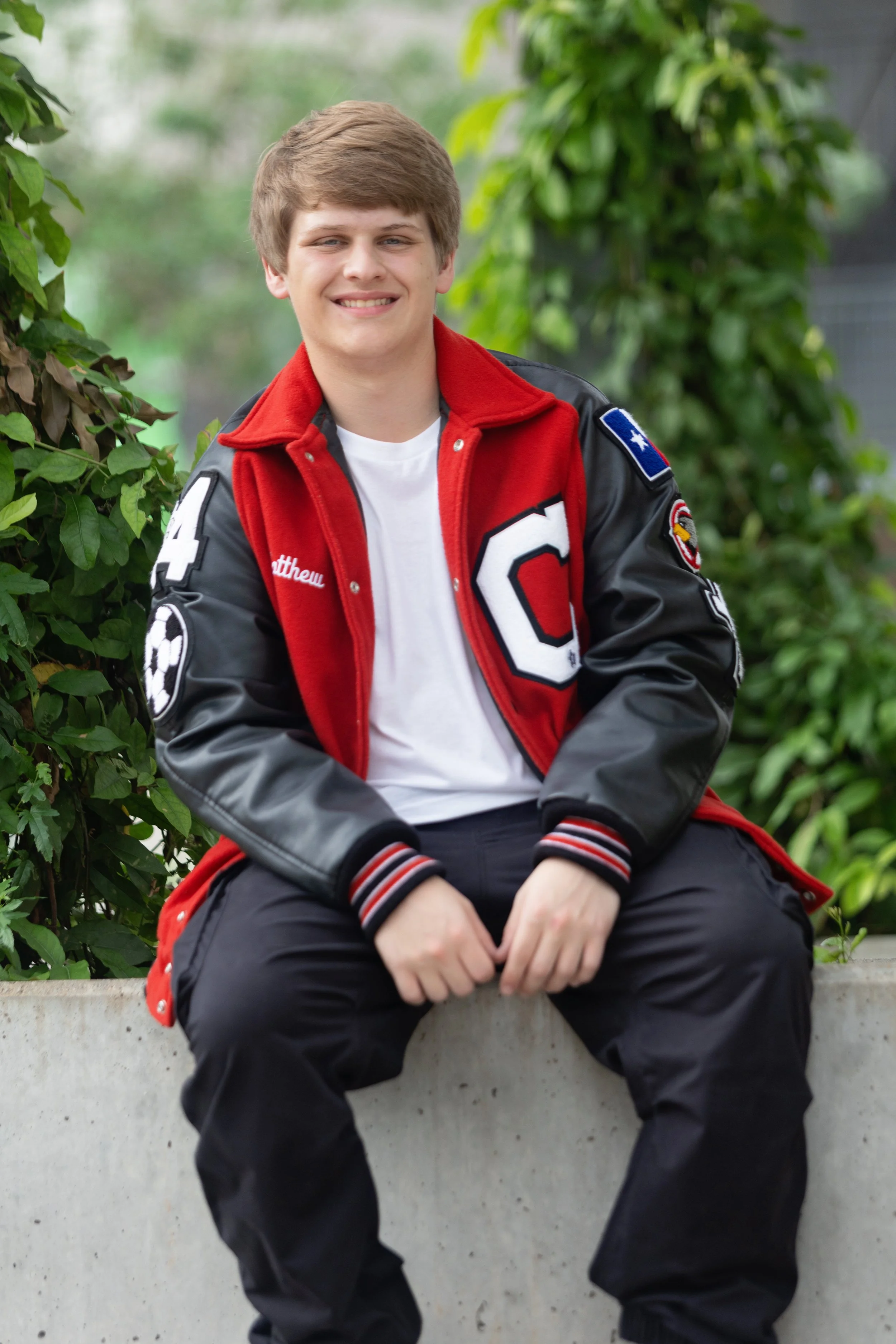 A young man with light brown hair, smiling, sitting on a concrete ledge outdoors surrounded by green foliage, wearing a red and black varsity jacket with patches and a white t-shirt.