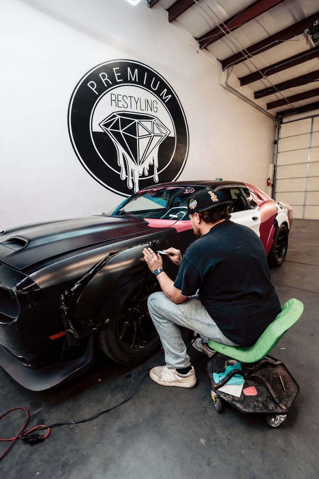 A man working on a black sports car with pink accents in an auto restoration shop. The wall features a large sign with a diamond and the words 'Premium Restyling.'