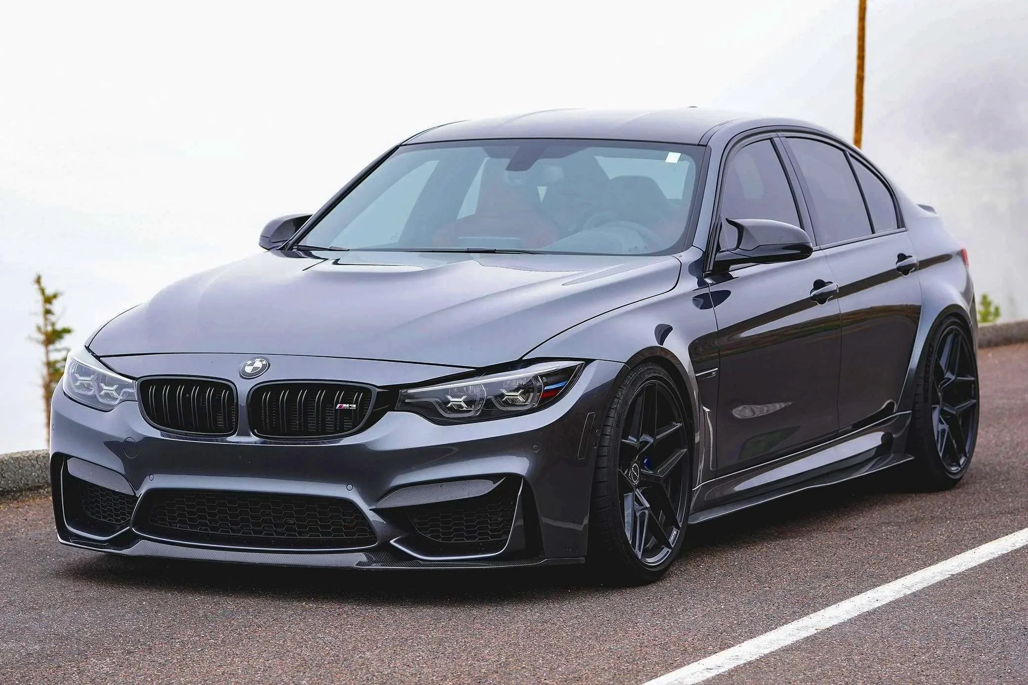 A sleek, dark gray BMW M3 sedan parked on a road near a body of water.