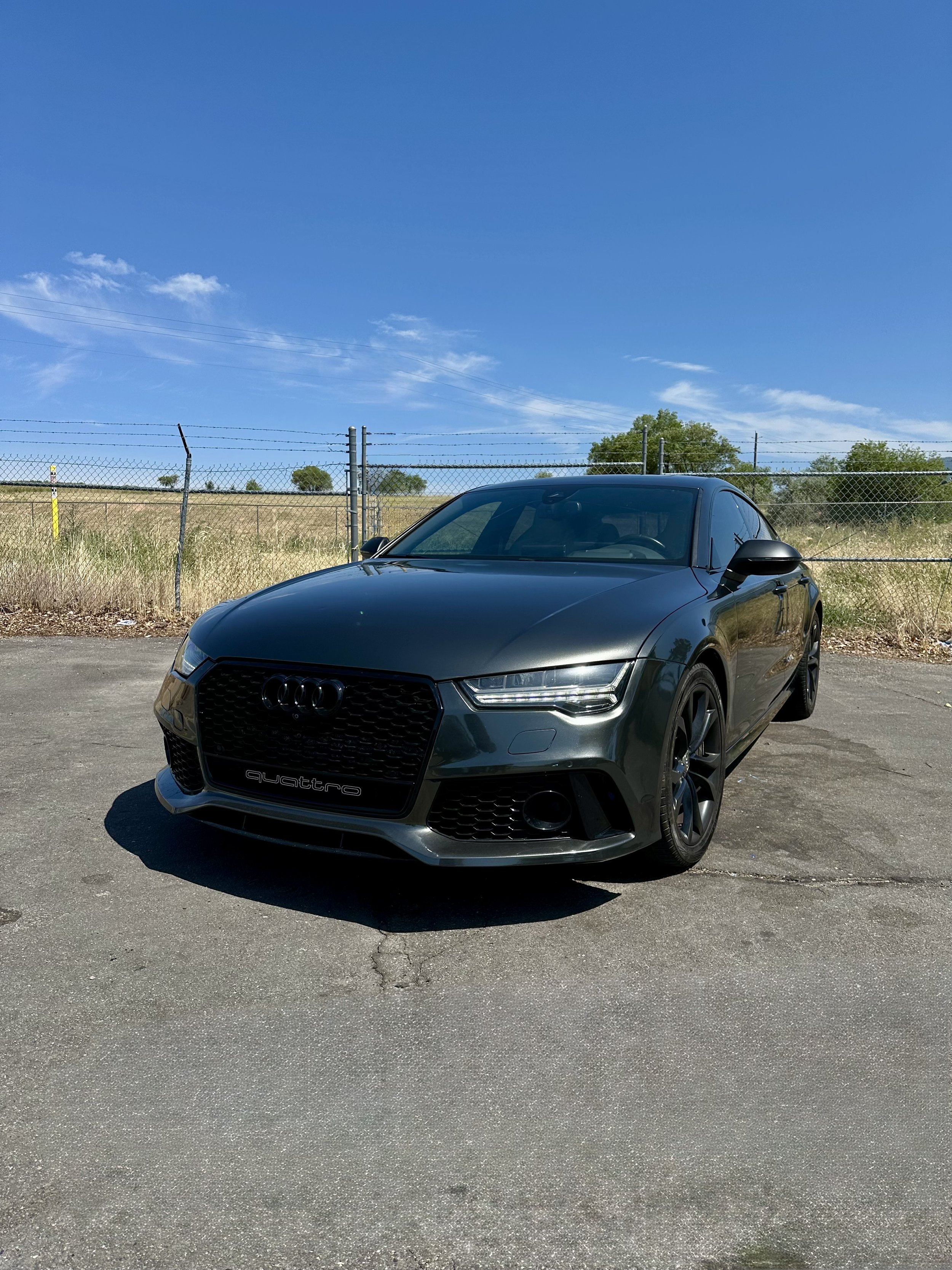 Black Audi Quattro parked on an asphalt lot with a chain-link fence and open field in the background under a clear blue sky.
