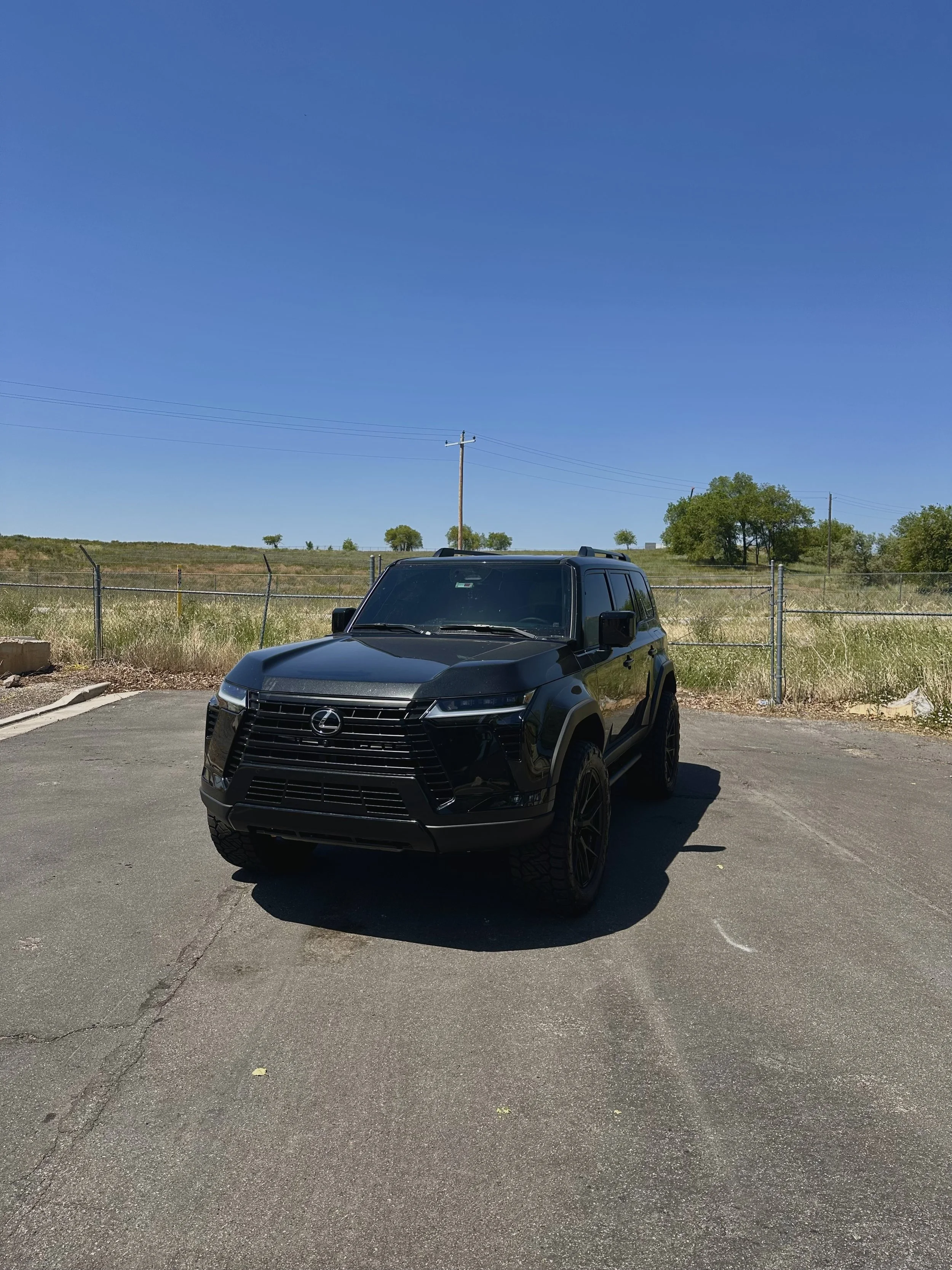 Black Lexus SUV parked on an asphalt lot under a clear blue sky, with a grassy field and a chain-link fence in the background.