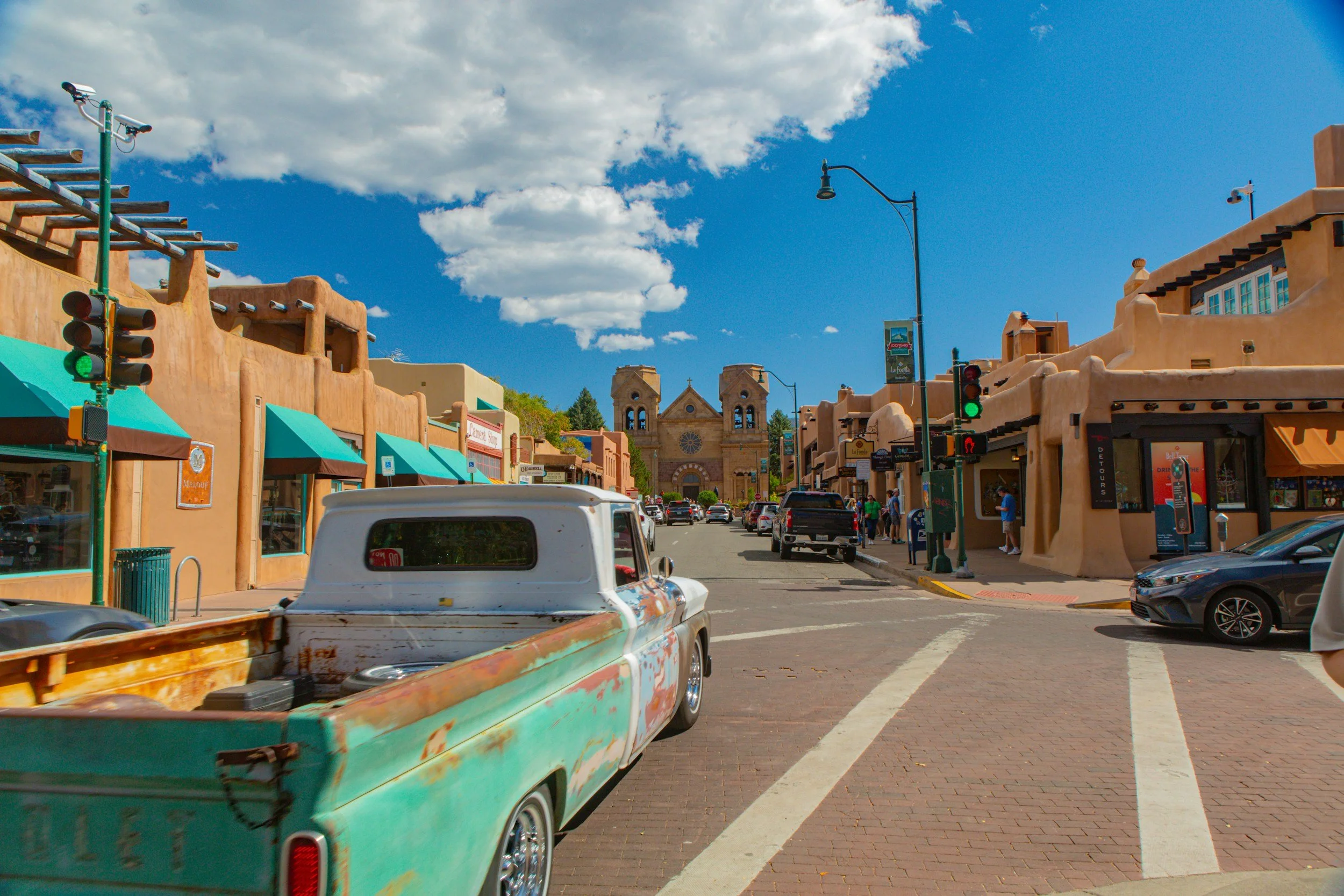 Downtown scene with colorful adobe-style buildings, parked cars, and a church with twin towers in the background under a partly cloudy sky.