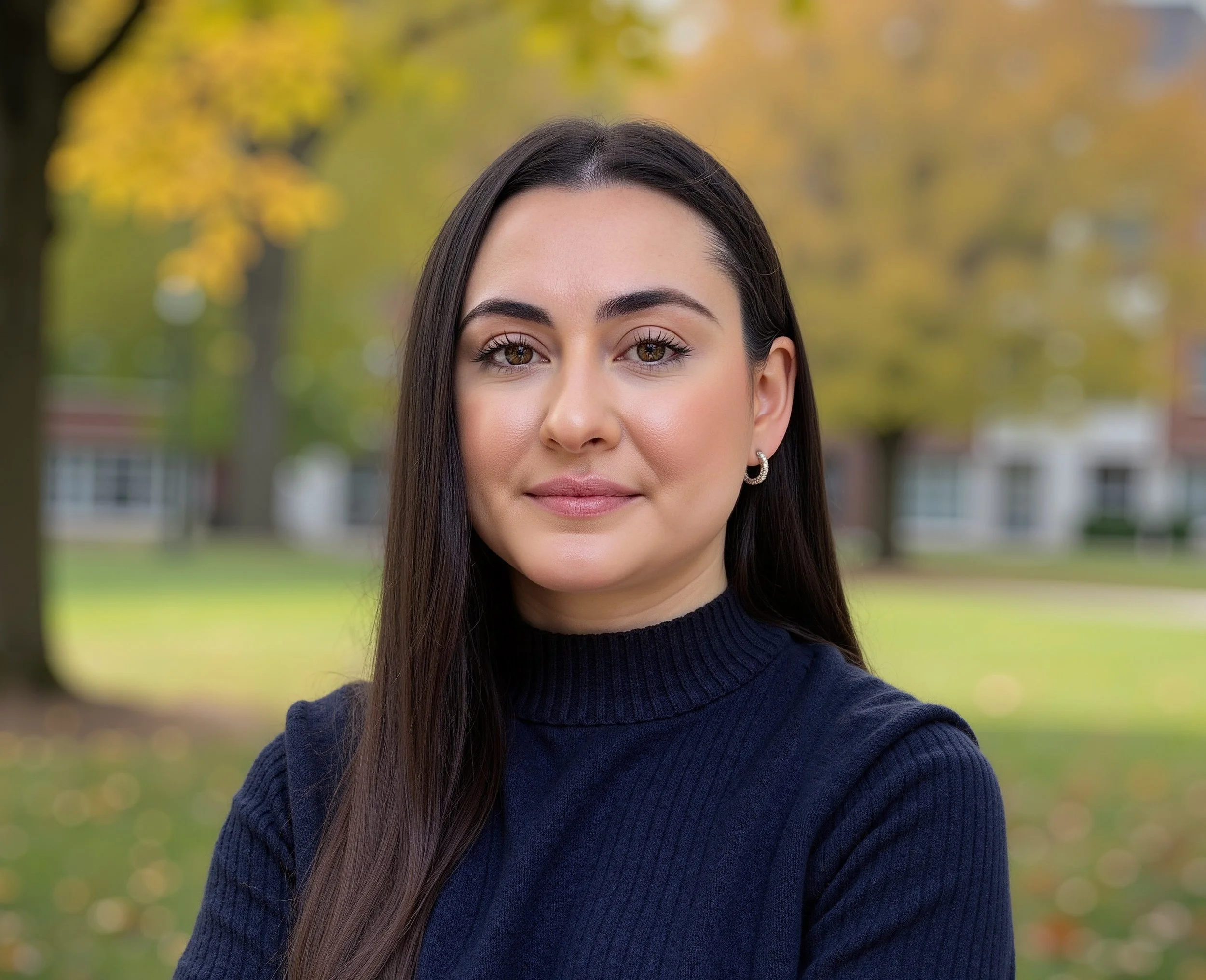 A woman with long dark hair, wearing a navy blue sweater and silver hoop earrings, standing outdoors in front of trees with yellow and green leaves in autumn.