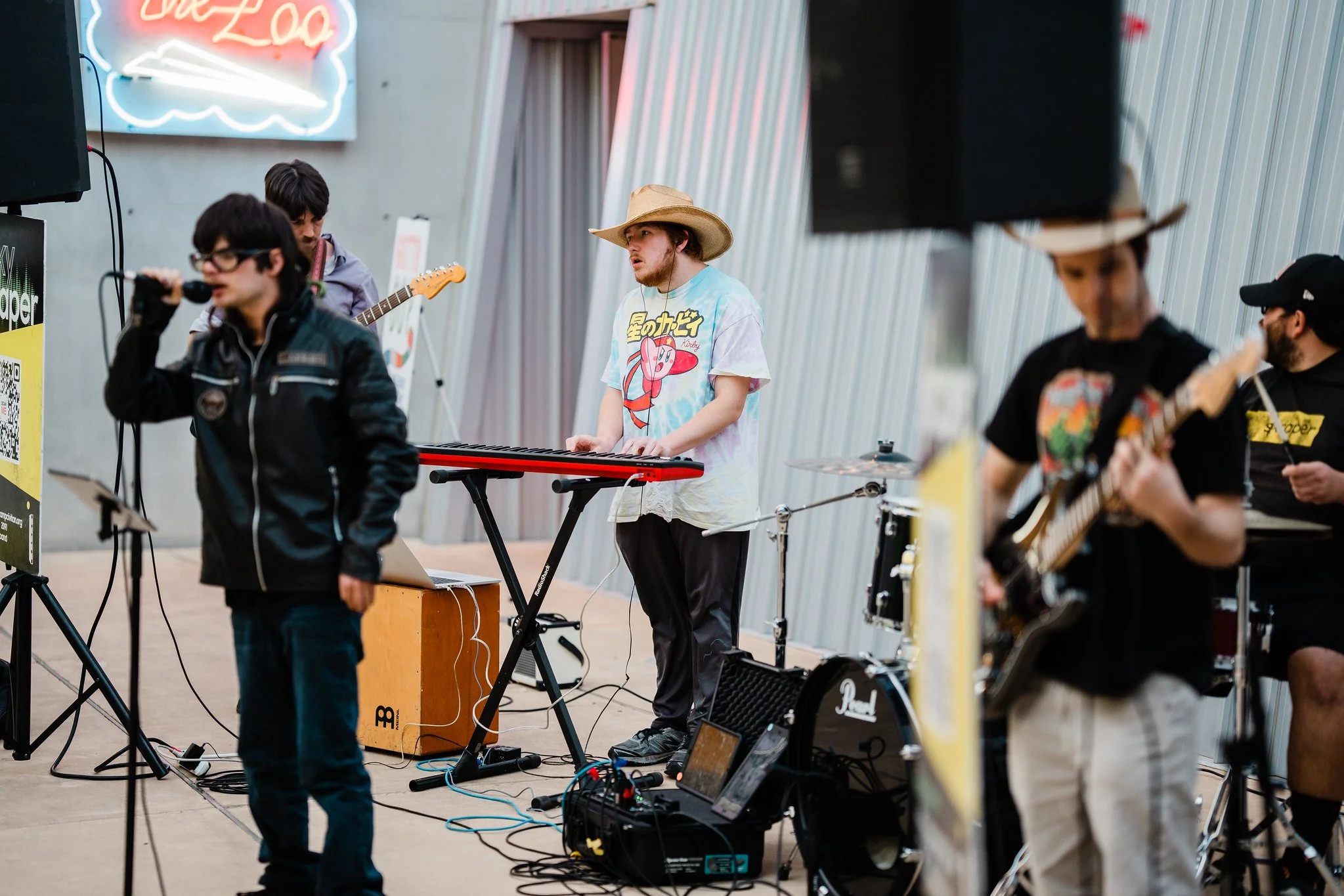 A band performing indoors with a singer, a keyboardist wearing a cowboy hat, and guitar players. Neon sign in the background.
