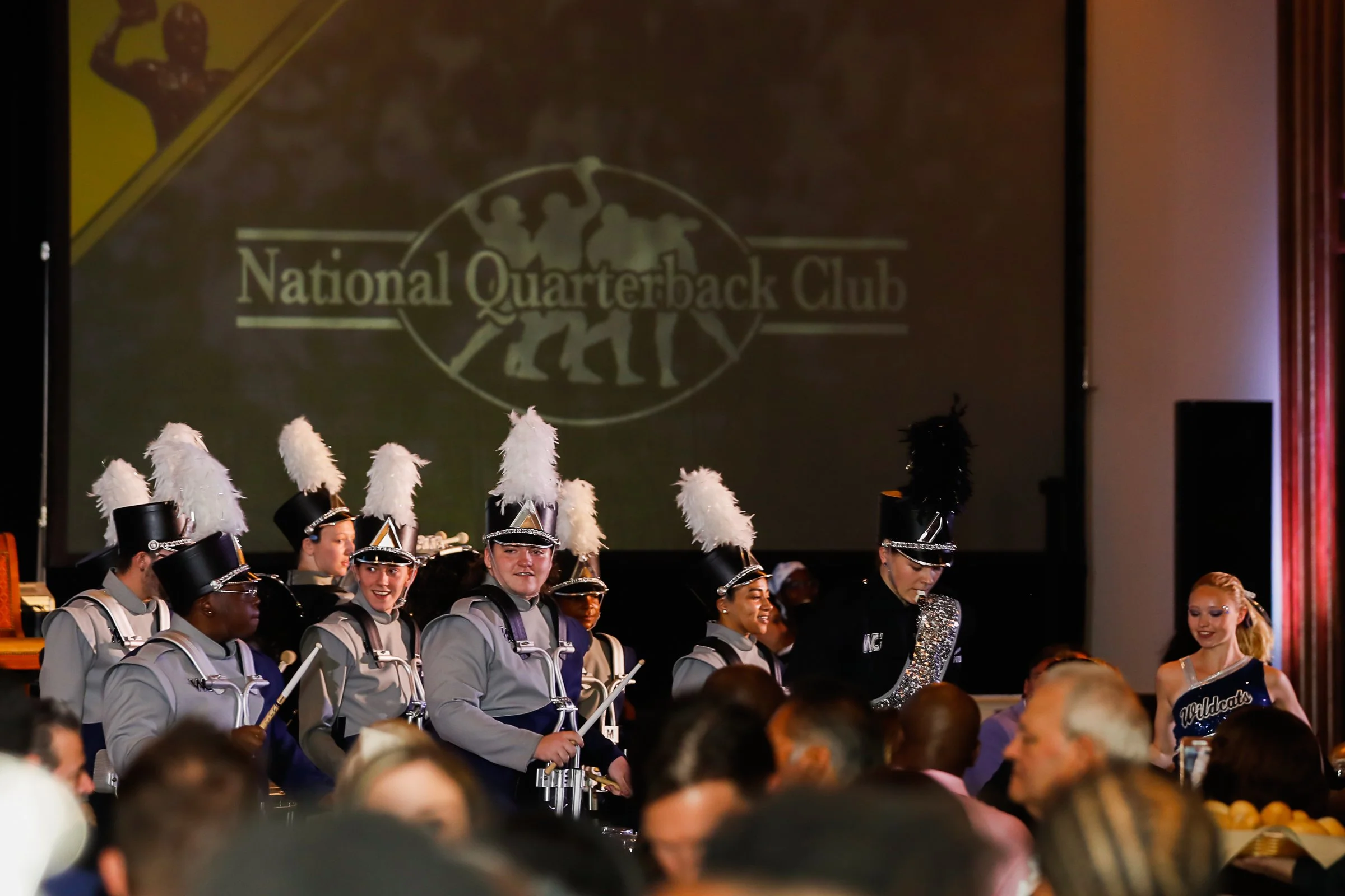 A group of marching band members dressed in grey uniforms with tall hats featuring white plumes, performing on stage at a National Quarterback Club event.