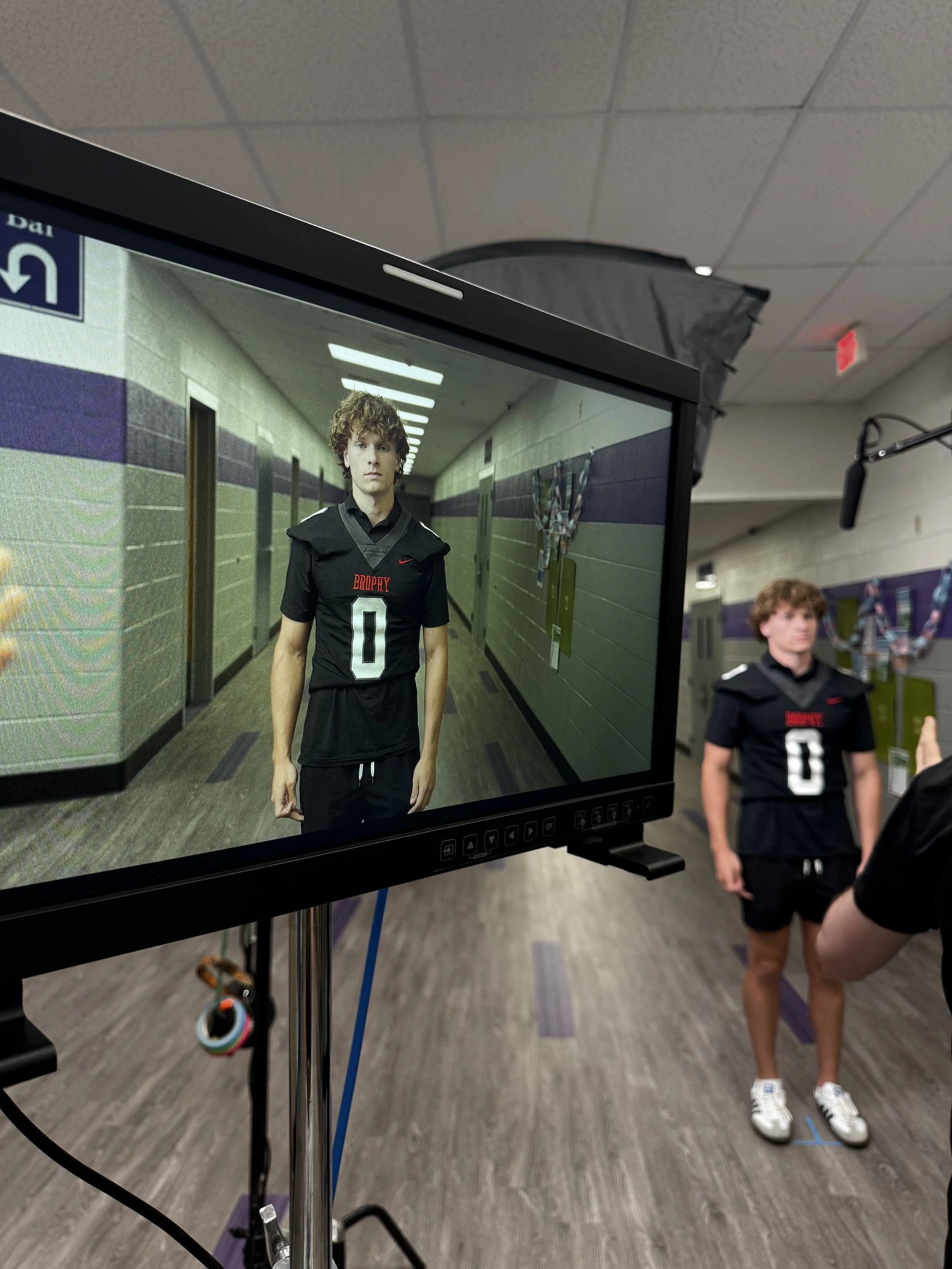 A young man in a black sports uniform standing in a school hallway during a photoshoot, as seen on a monitor. The hallway has gray walls with purple accents and several closed doors.