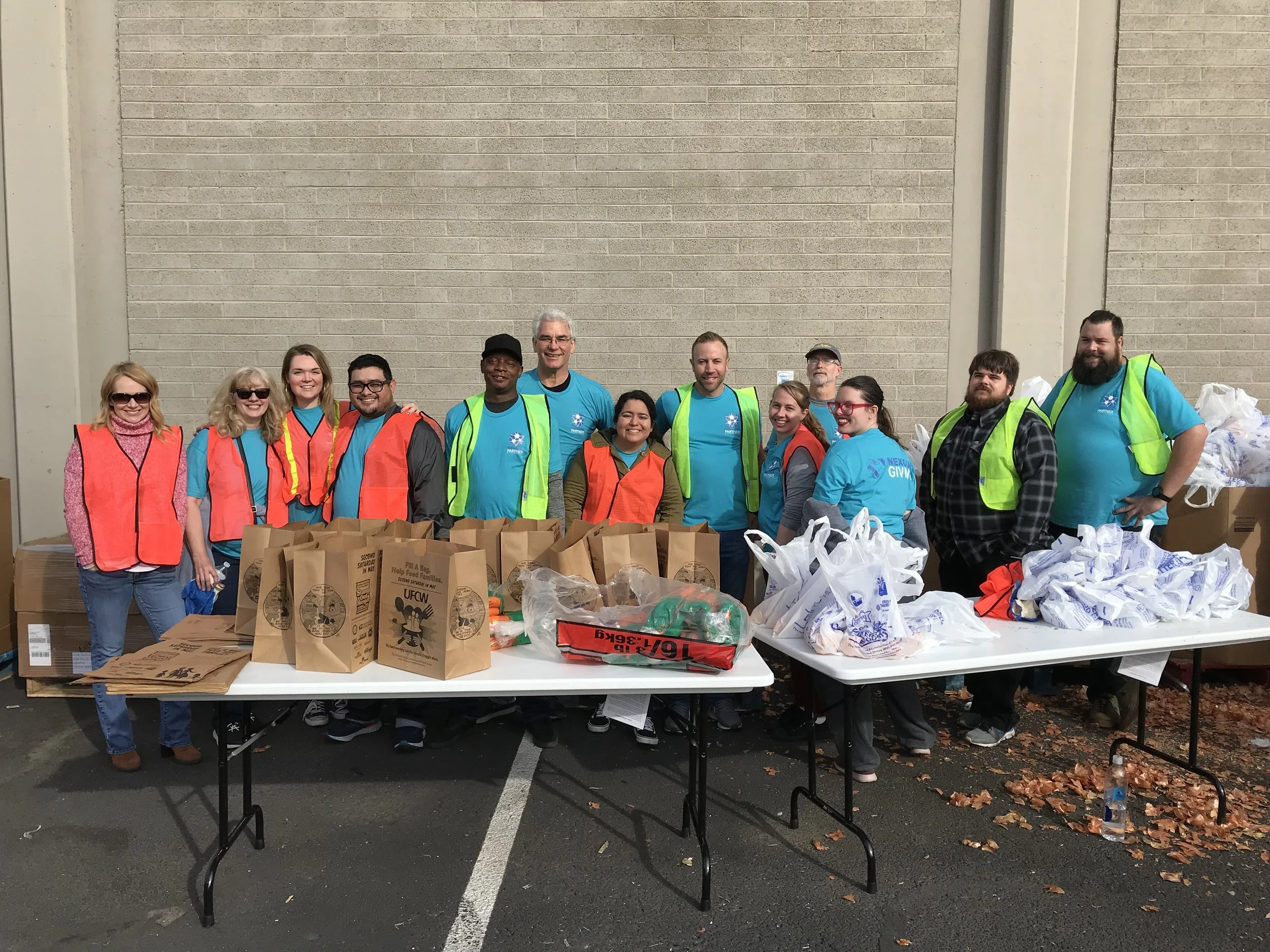 Group of volunteers wearing orange and green safety vests standing behind tables with grocery bags and supplies outdoors.