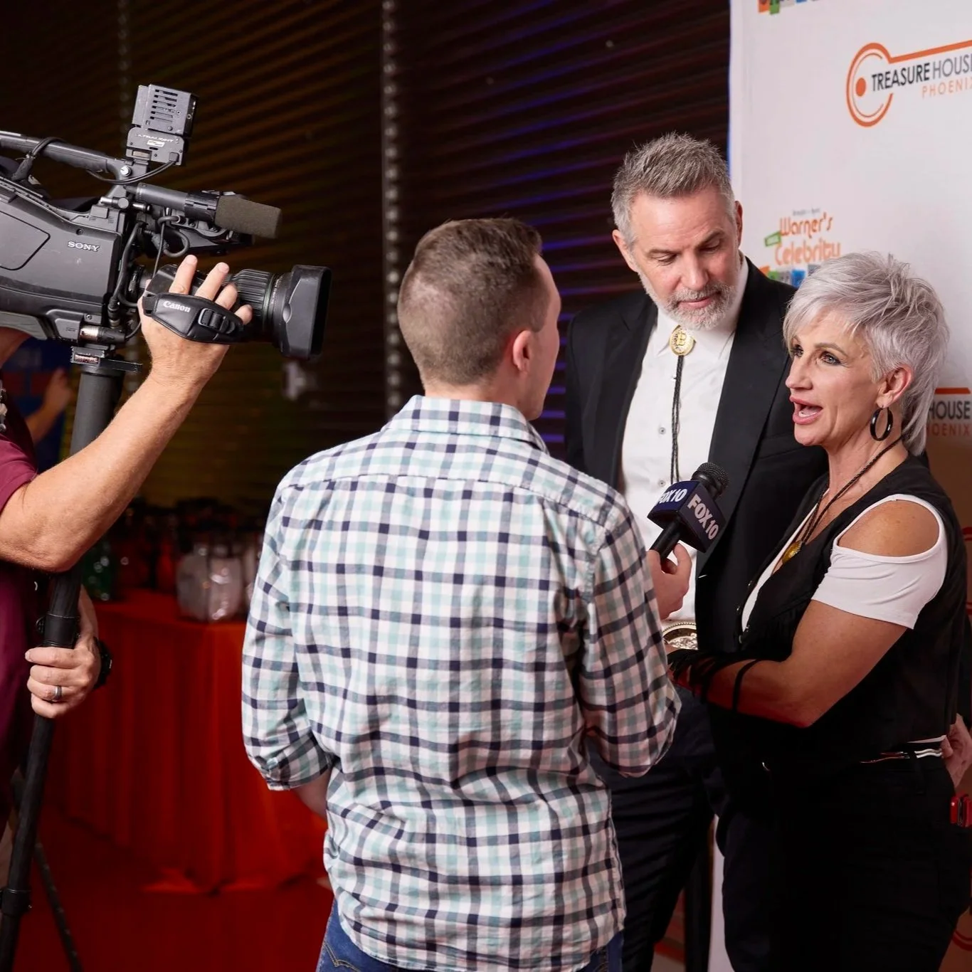 A woman being interviewed by a man holding a microphone, with a cameraman filming the scene. A man with gray hair and beard wearing a black blazer stands next to her. They are at a public event with a banner in the background.