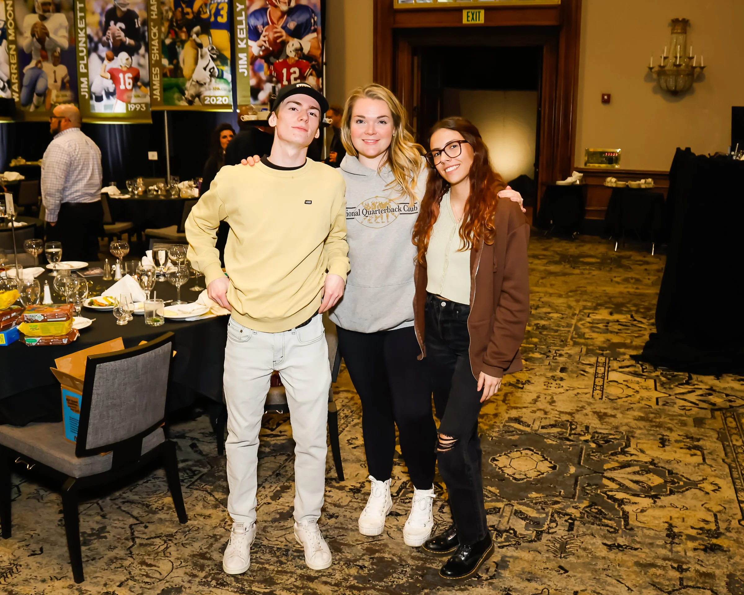 Three young adults, two women and one man, smiling and posing together in a banquet hall decorated with football banners. They are dressed casually, standing close with their arms around each other.