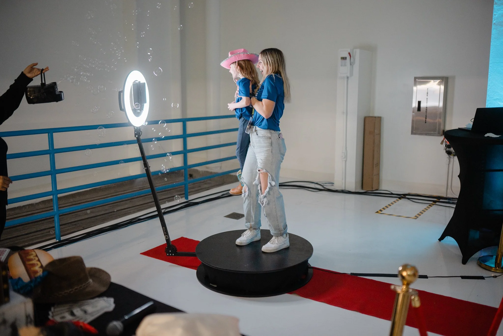 Two women standing on a circular platform, one holding a young girl with a pink hat, as a photographer takes their picture in a studio with a red carpet and blue railing.
