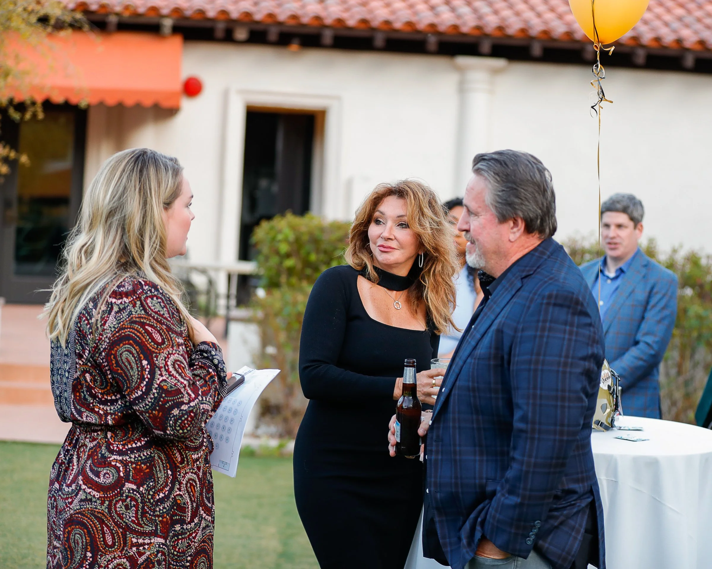 People socializing at an outdoor gathering, with a woman holding a clipboard and a woman with red hair and a man with gray hair holding a beer, engaging in conversation near a decorated table with balloons.