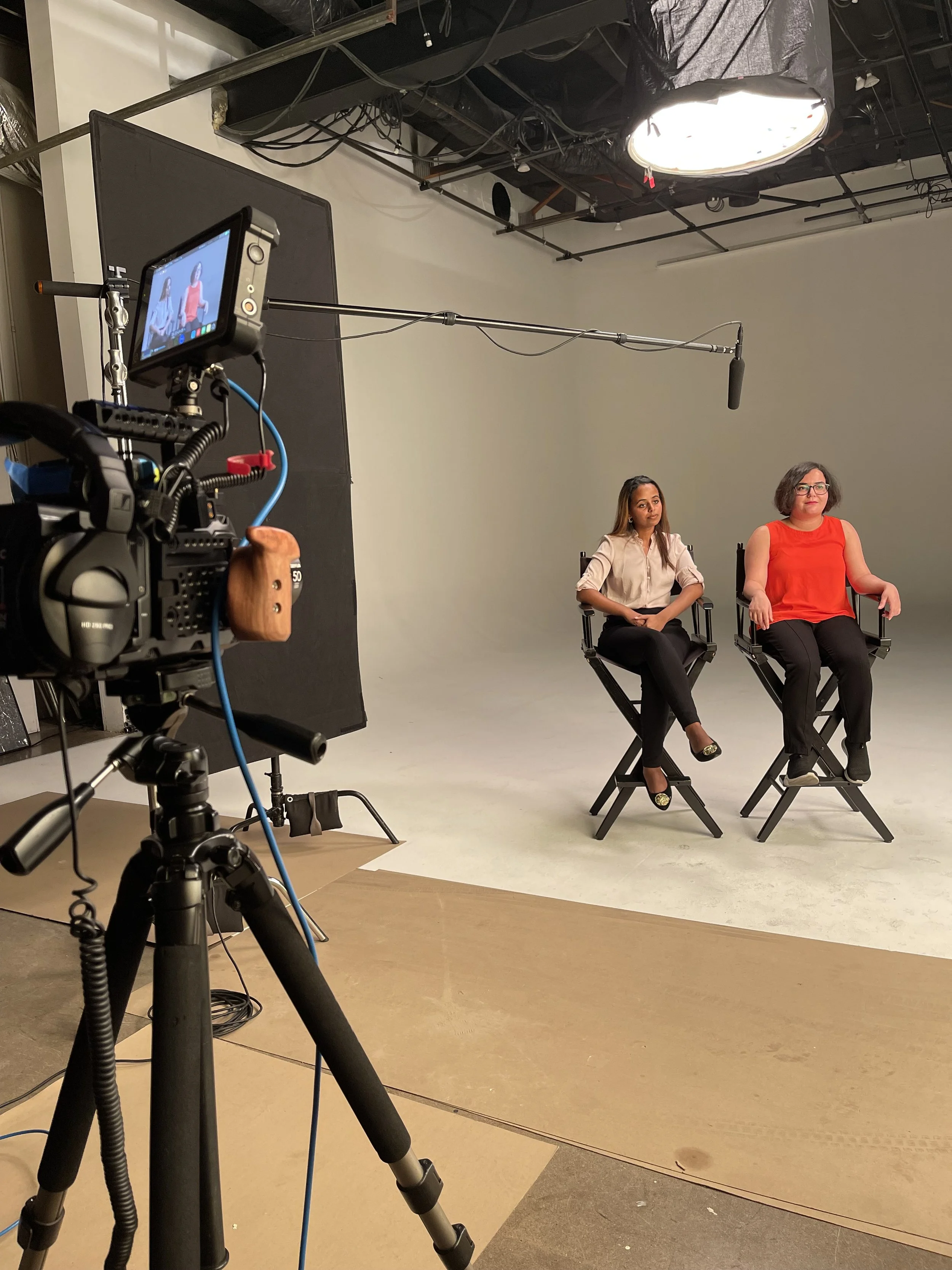 Two women sitting on director's chairs in front of a blank wall during a video shoot, with professional camera equipment set up in a studio.