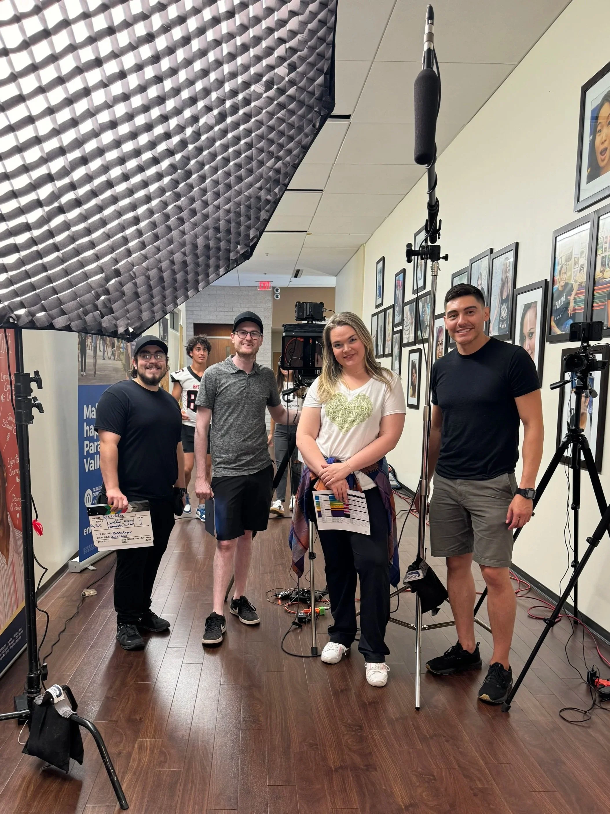 Group of five people filming in an indoor hallway decorated with framed photographs, with professional lighting and camera equipment around them, including a large softbox light and a boom microphone.