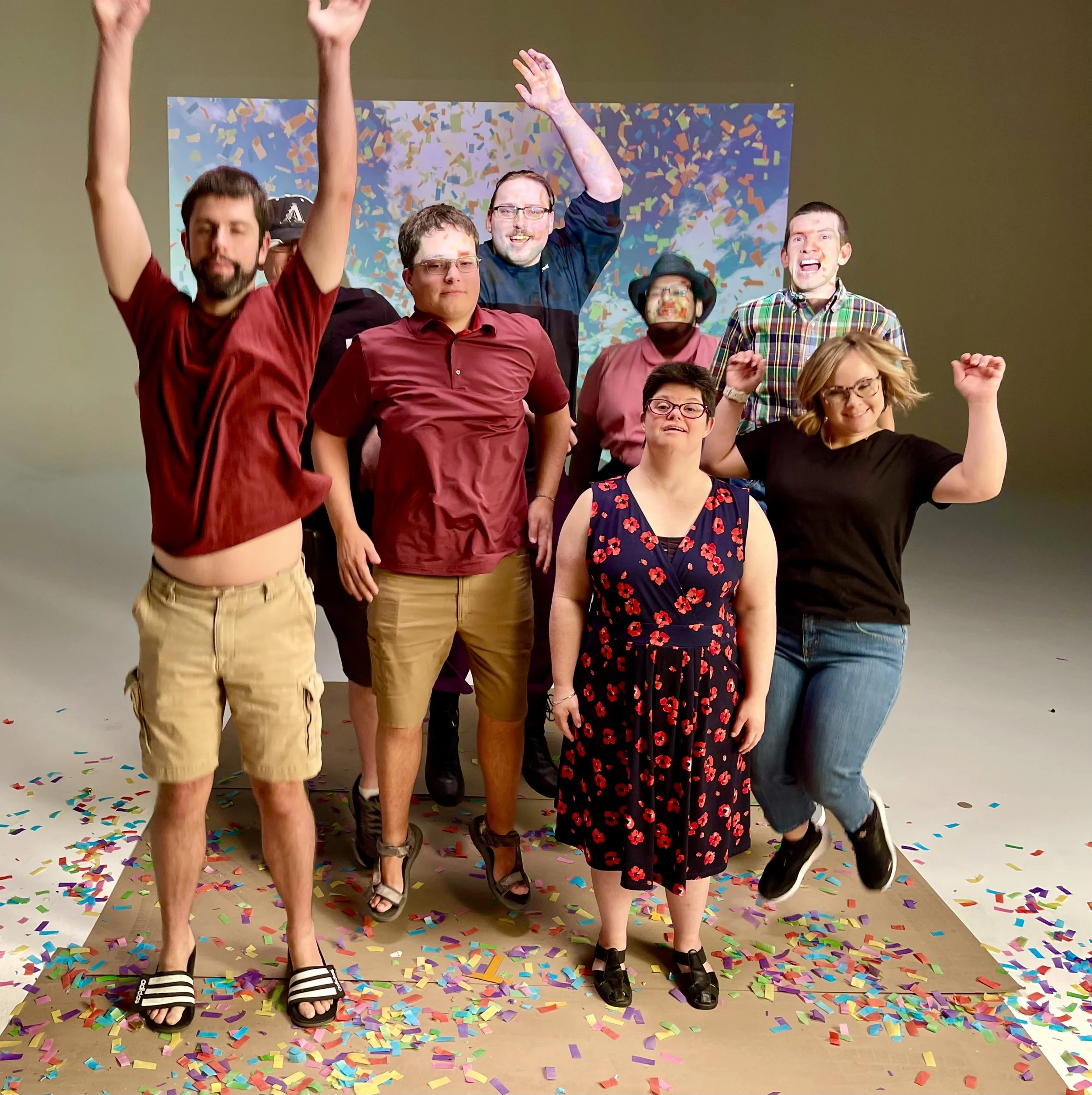 Group of eight people celebrating with colorful confetti on the floor, some jumping, some standing, in a studio with a plain background.