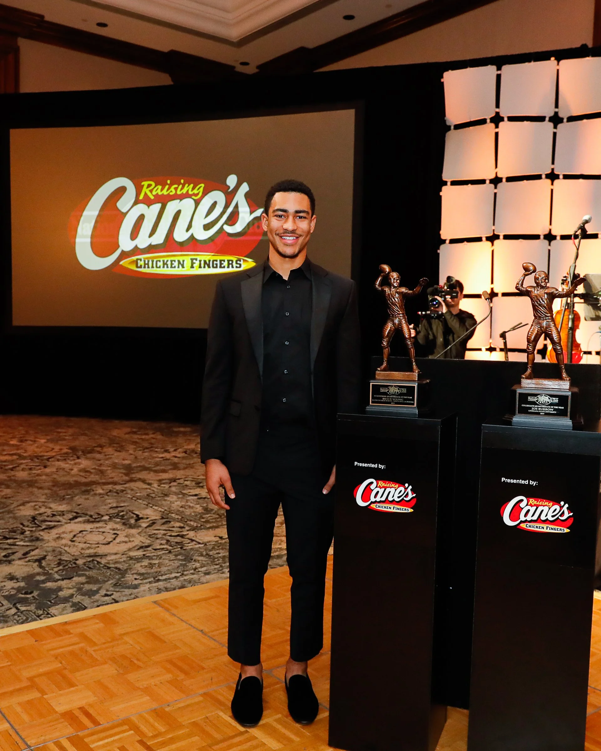 Young man in a black suit standing next to two trophies at an event with a large screen displaying 'Raising Cane's Chicken Fingers' in the background.