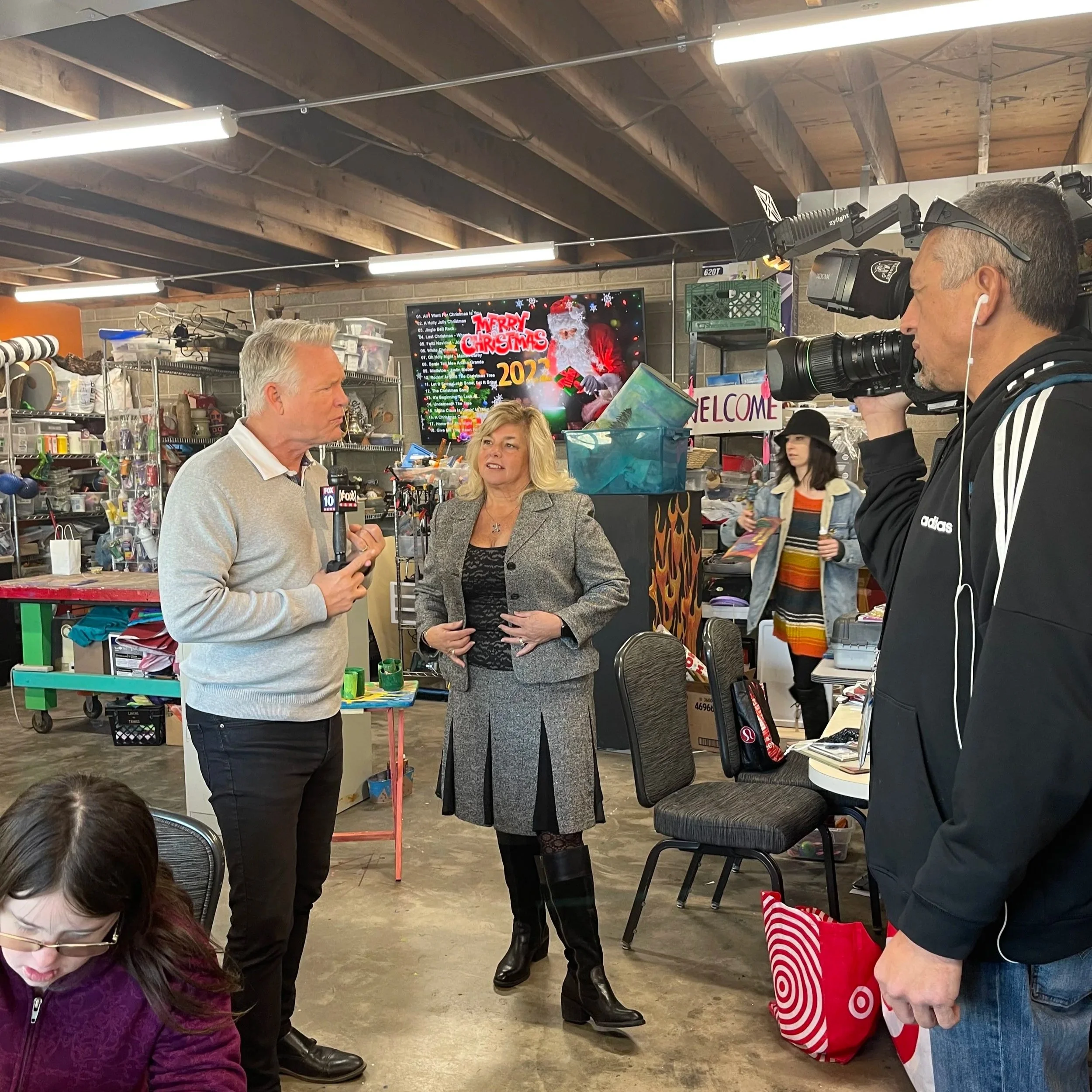 News crew filming a woman being interviewed in a cluttered room with shelves and Christmas decorations on the screen in the background.