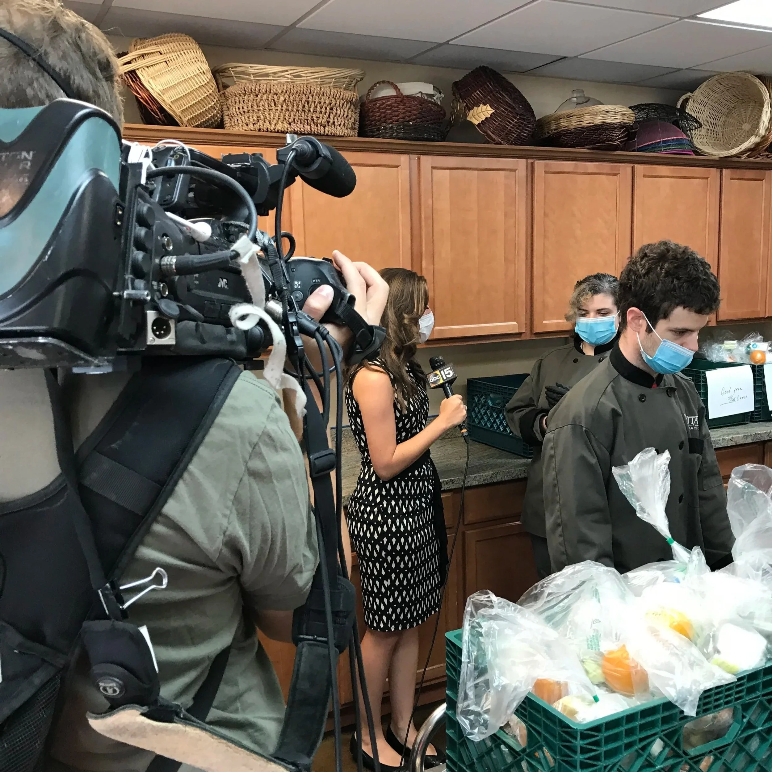 A news reporter holding a microphone interviews a worker in a kitchen or food distribution setting, with a cameraman filming the scene. The worker and others are wearing masks, and there are baskets and food items on a counter.