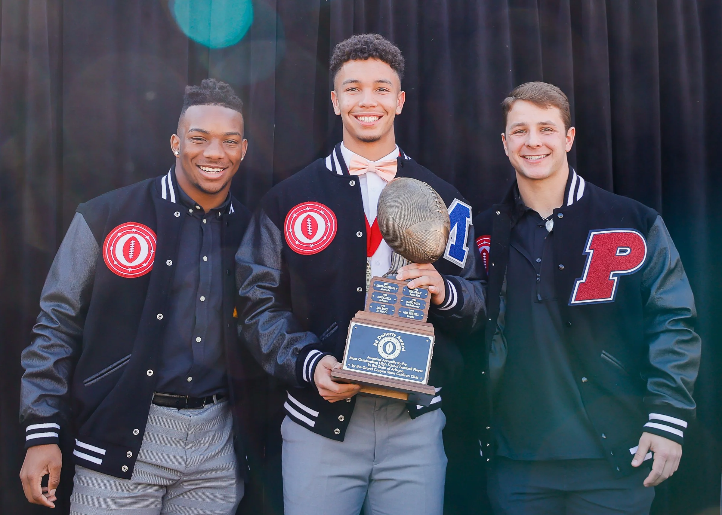 Three young men in varsity jackets standing together, with the man in the middle holding a trophy shaped like a football, smiling for a photo.