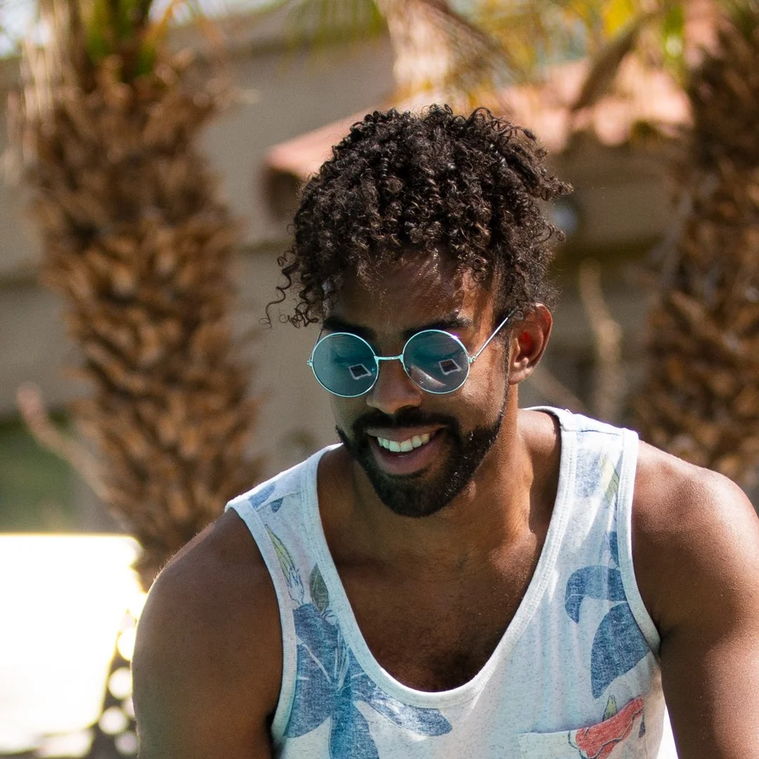 A smiling man with curly hair and sunglasses, wearing a sleeveless shirt, outdoors with palm trees in the background.