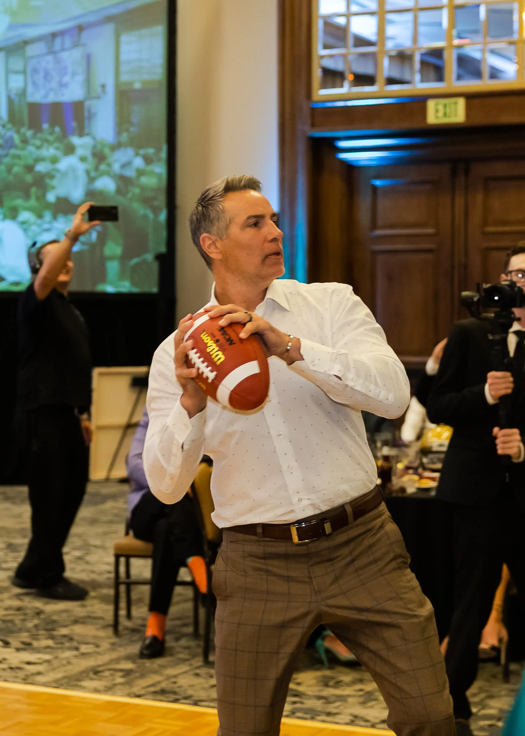 A man in a white shirt and brown plaid pants holding a football at an indoor event, with other people taking photos and a large projection screen in the background.